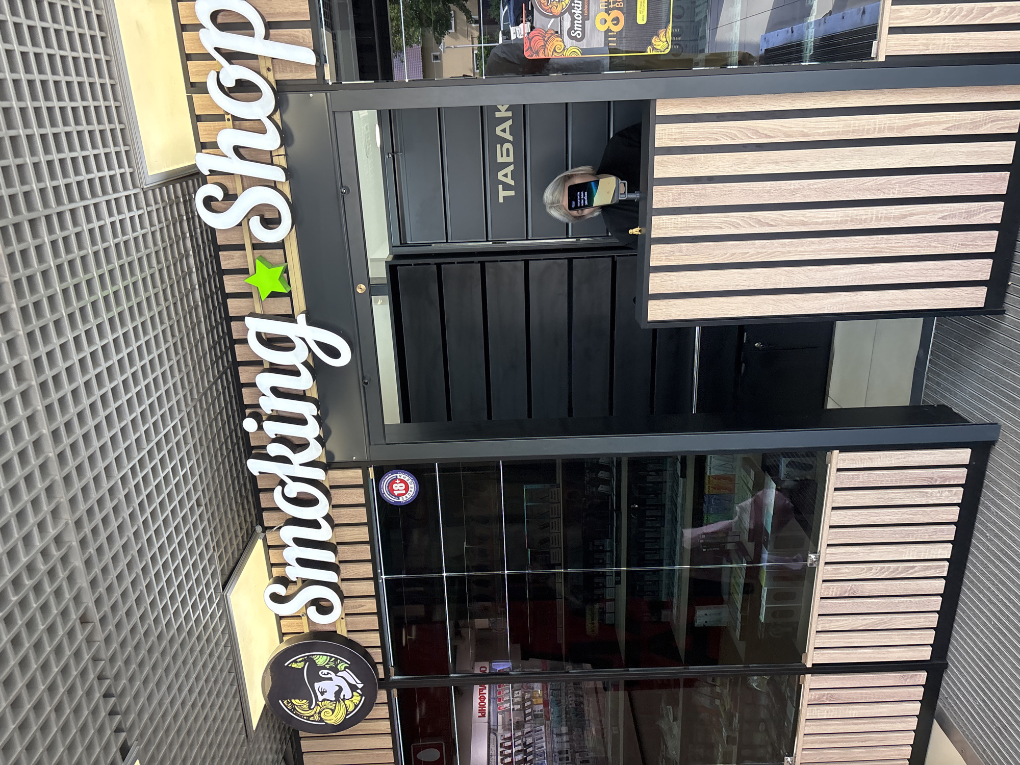 The image shows the storefront of a "Smoking Shop" located in Gorki-2, Russia. The shop's name is prominently displayed in large, white, cursive lettering above the entrance. To the left of the name is a circular sign featuring a stylized black and white illustration of a man wearing a top hat, with yellow and green accents. A green star is positioned between the words "Smoking" and "Shop". The shop's facade consists of large glass display cases showcasing various tobacco products and accessories. The entrance area is dark, with horizontal black panels above the counter. A person with blonde hair is partially visible behind the counter, holding a smartphone that obscures their face, displaying a message in Cyrillic. To the right of the entrance, another sign for "Smoking Shop" is visible, along with what appears to be an 18+ age restriction symbol. The exterior of the shop features wooden paneling and is situated in what appears to be a commercial or shopping area, with a street and buildings visible in the background through the glass. The ground in front of the shop is covered with metal grating.
