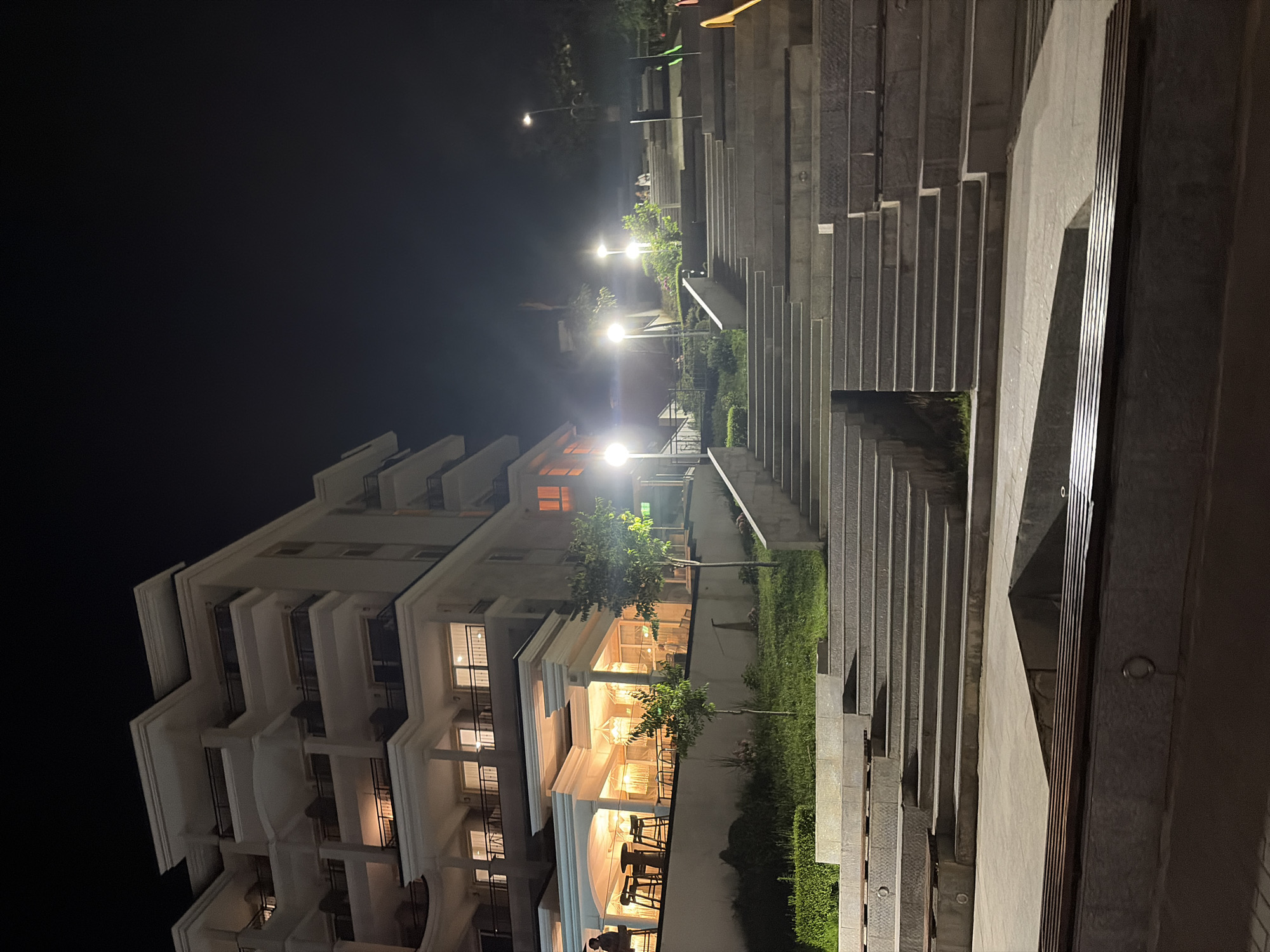 The image depicts a modern, multi-story building at night, likely a hotel or residential complex, with illuminated balconies and windows. A wide set of stone stairs with integrated lighting leads up from a paved area with wooden benches towards the building. The building's facade is light-colored, with several levels featuring balconies and decorative columns. Below the building, an outdoor seating area, possibly a restaurant or cafe, is visible with tables and chairs illuminated by warm, ambient lighting. Small trees and manicured greenery are present on the slopes alongside the stairs. Streetlights cast a glow on the scene, illuminating pathways and the surrounding environment. A few individuals are discernible sitting on the stairs in the mid-ground. The overall atmosphere is serene and inviting, suggesting a tranquil evening setting in Obzor, Bulgaria.