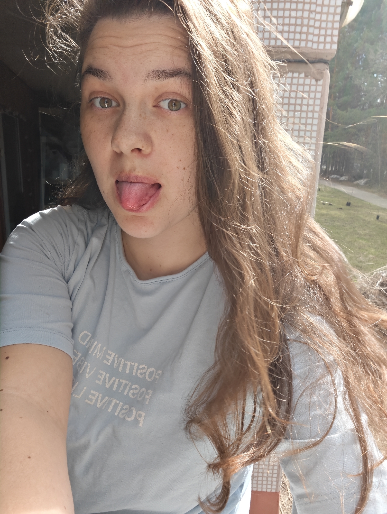 A young woman with light brown, wavy hair and freckles on her face is captured in a bright, outdoor selfie. She looks directly at the camera with a playful expression, sticking her tongue out. Her eyes are light green or hazel. She is wearing a light blue t-shirt, on which the mirrored text "POSITIVE VIBE" is visible multiple times.
The image is taken during daytime, with strong sunlight illuminating her hair, creating a bright halo effect and suggesting a sunny weather. The background features elements of both urban and natural environments. To the left and upper right, a light-colored building facade with a distinctive square tiled pattern is visible, likely part of a balcony or exterior wall. Beyond this structure, a landscape of tall trees, green grass, and a winding path or road can be seen, hinting at an early spring or late autumn setting given the sparse ground cover in some areas. The overall mood is casual and cheerful. The location context suggests this scene is in Chaikovsky, Russia.