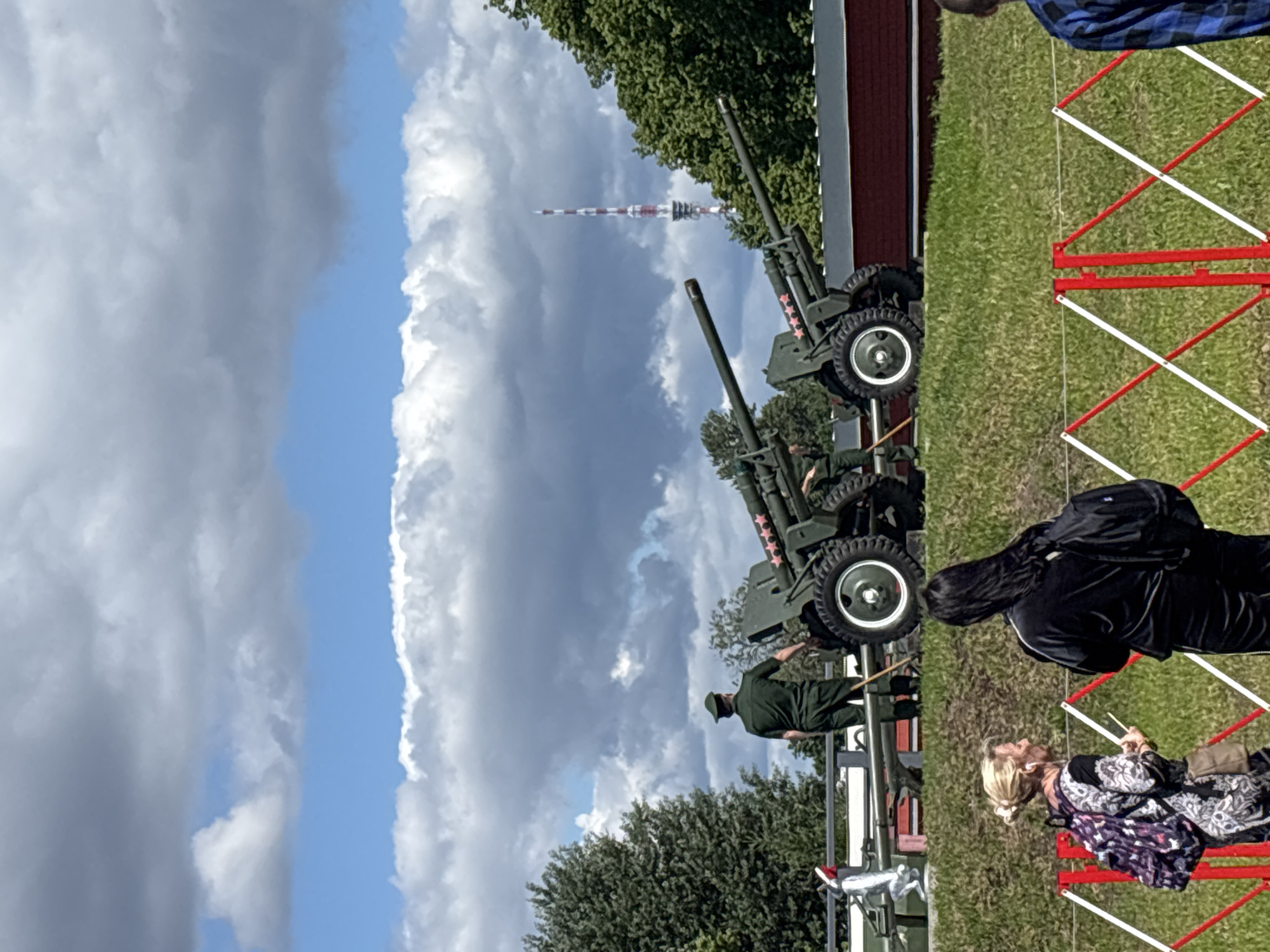The image captures an outdoor scene with a blue sky featuring scattered white and gray clouds. In the middle ground, two green artillery cannons are positioned on a grassy area. A person in a green military uniform stands near the front cannon, facing away from the camera. To the right of the cannons, a tall, red and white striped tower, likely a radio or television tower, is visible behind a line of green trees. In the foreground, two individuals are observed from behind, standing on a grassy slope separated by a red and white striped barrier. The person on the left has blonde hair pulled back and is wearing a patterned dress and a backpack, while the person on the right has long dark hair and is wearing a dark tracksuit and a backpack. A red building is situated behind the cannons. The overall setting appears to be a park or historical site.