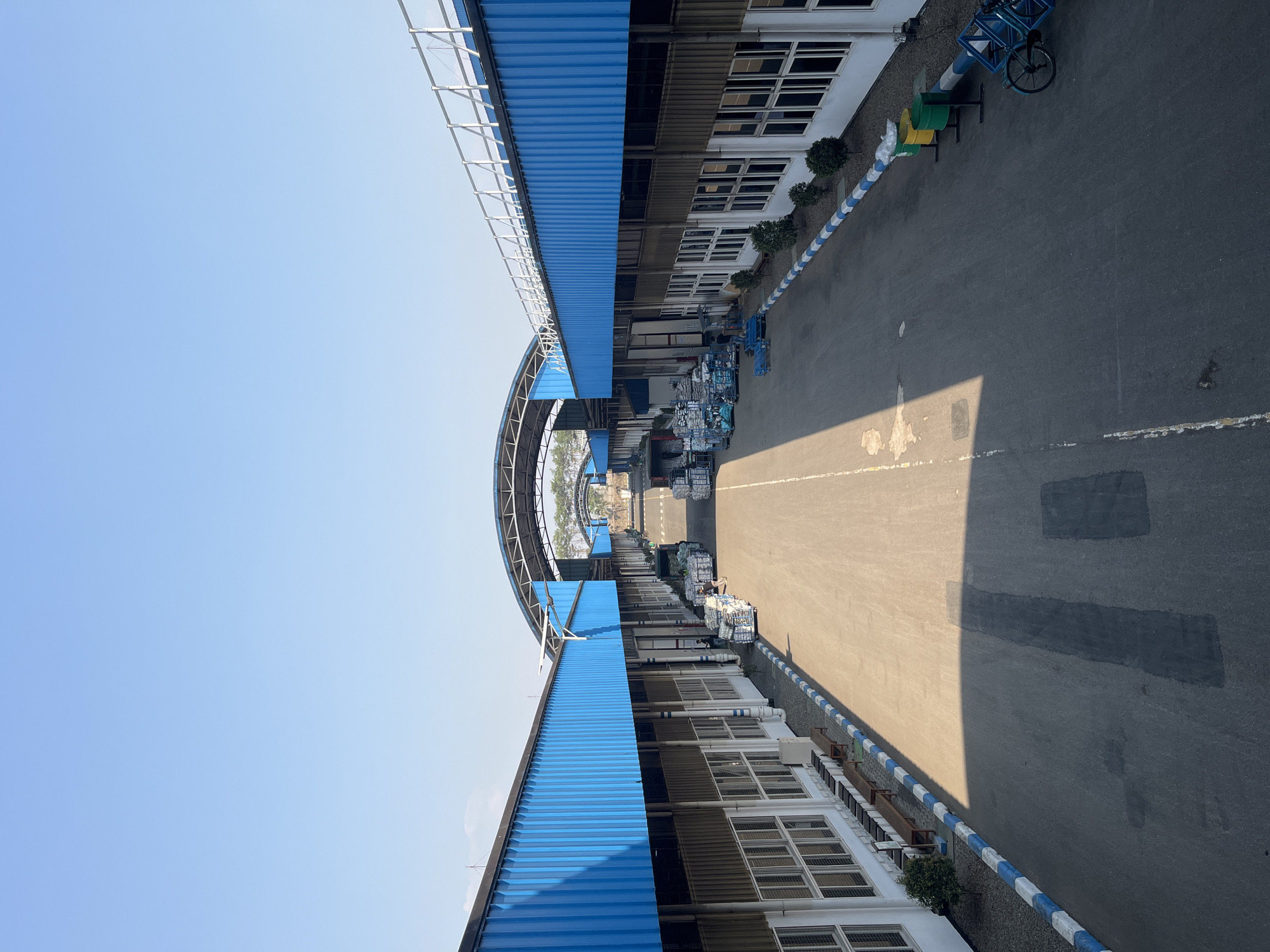 The image depicts a view down a long, paved pathway between two parallel industrial buildings with blue corrugated metal roofs. The pathway is lined with benches on the left and trash receptacles and a blue bicycle on the right. Several stacks of goods, possibly merchandise or supplies, are visible along both sides of the pathway, with some being moved by people. The buildings have numerous windows and are supported by a structure that creates arched overhangs in the distance. The sky above is clear and blue, indicating an outdoor daytime scene in Pelang, Indonesia.