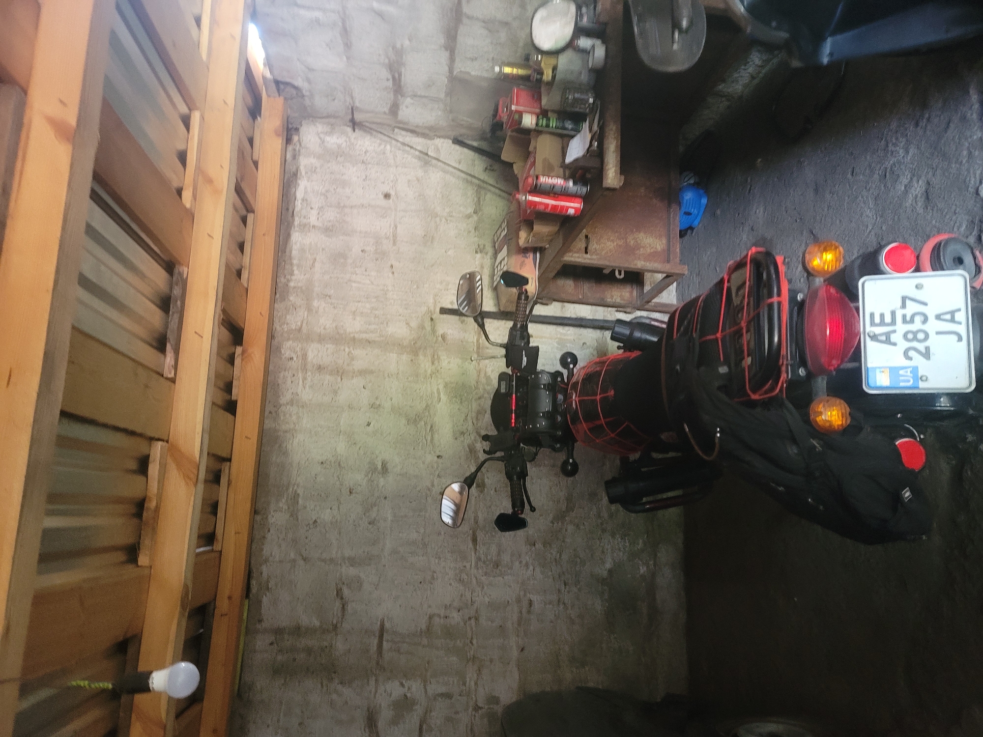In the city of Vovkivka, Ukraine, a motorcycle is parked inside a dimly lit garage. The garage has rough concrete walls and a wooden plank ceiling. A single light bulb hangs from the ceiling, illuminating the scene. The motorcycle is black with red accents and has saddlebags attached. A license plate with the characters "AE 2857 JA" is visible on the rear of the motorcycle. To the right of the motorcycle, there is a metal workbench with various items on it, including cans, boxes, and tools. The ground is made of concrete.