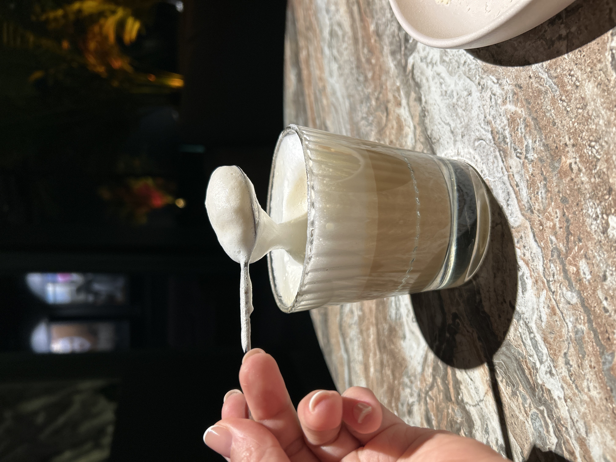A human hand with manicured nails and white French tips holds a small, silver-colored spoon, lifting a portion of white foam from a drink. The spoon is positioned directly above a clear, ribbed glass containing a layered beverage: a light brown liquid at the bottom topped with a thick layer of white foam, some of which has spilled over the rim. The glass and a partial view of a light-colored, possibly cream, ceramic dish with crumbs rest on a mottled brown and grey surface, likely a stone tabletop. The background is dark and out of focus, featuring blurred warm light sources and indistinct shapes. The scene is set indoors, likely within a cafe or restaurant in the city of Chaikovsky, Russia.