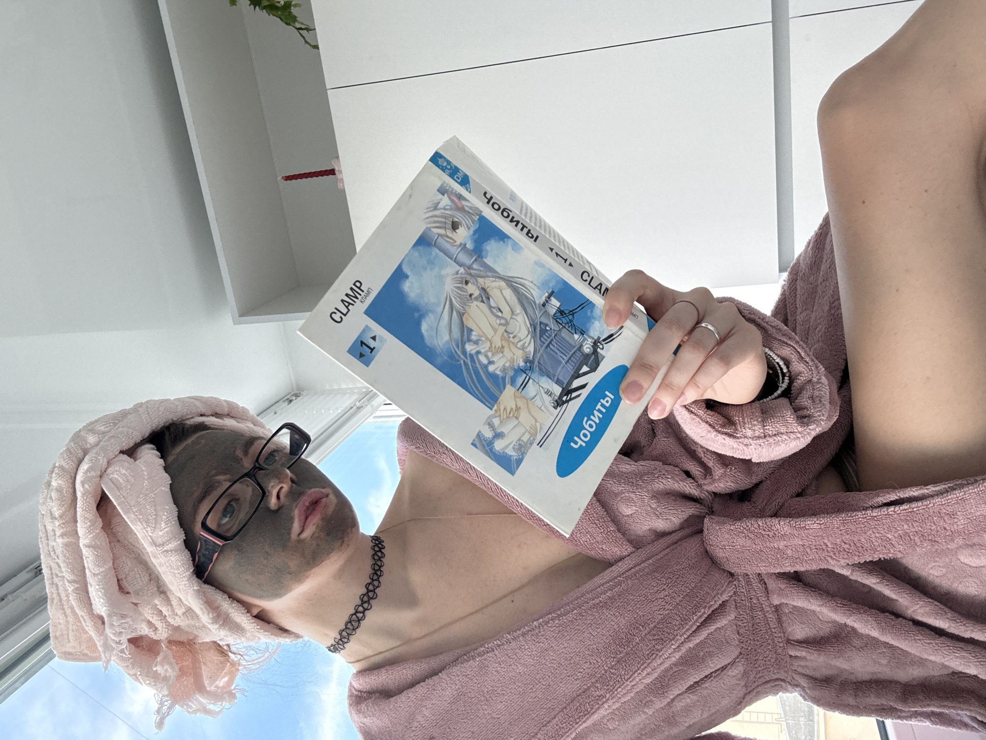 A young woman, wearing a pink bathrobe and towel turban, with a facial mask applied, sits by a window, reading a manga titled "CLAMP CHOBITS". She is wearing glasses and a choker necklace. The environment appears to be indoors, possibly a bedroom or bathroom, with a white shelf unit in the background.