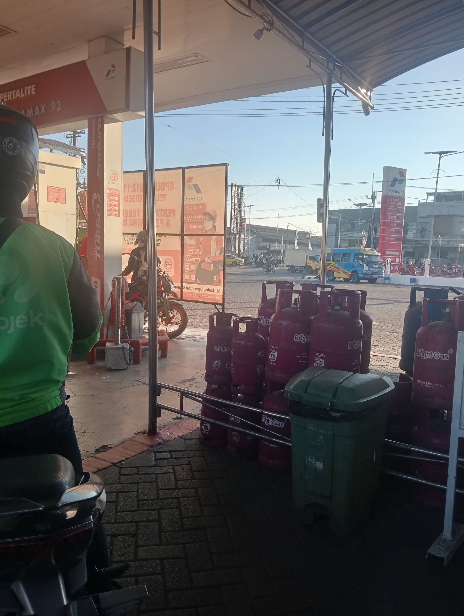 The image depicts a scene at a Pertamina gas station in Sawotratap, Indonesia. In the foreground, a person wearing a green jacket and a black helmet is seated on a motorcycle. To their right, a row of red and pink gas cylinders are stacked. A green trash can is positioned between the gas cylinders and the motorcycle. In the mid-ground, a person on a motorcycle is near a pump, and a Pertamina sign displays fuel prices. Beyond the gas station, traffic can be seen on a road, with cars and trucks, including a yellow and blue bus. The environment appears to be outdoors, with a clear blue sky and sunlight casting shadows.