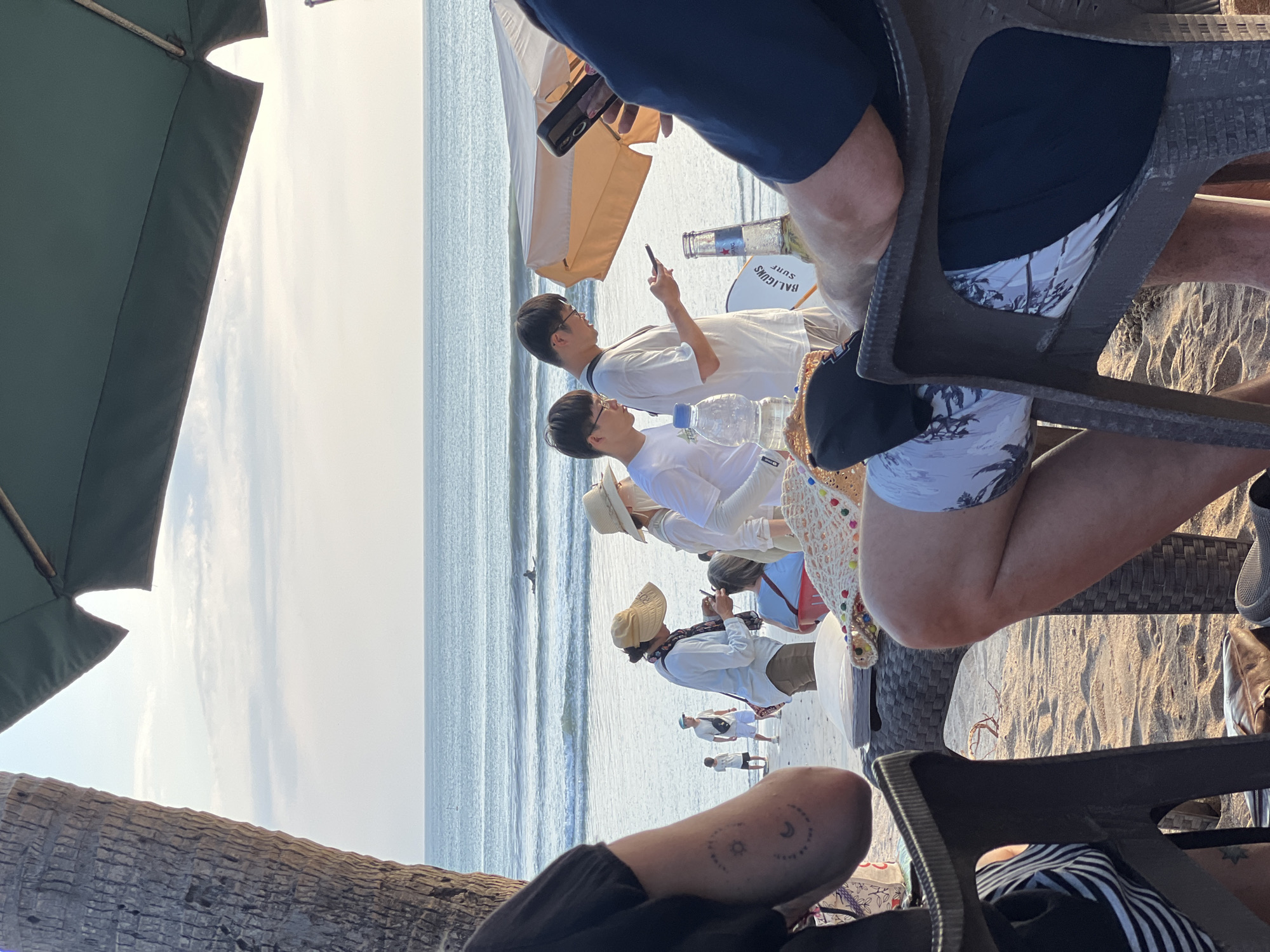 The image captures a beach scene in Kuta, Indonesia, likely during the late afternoon, given the soft, warm lighting. In the foreground, two figures are seated at a table. The person on the left is partially visible, wearing a black t-shirt and striped shorts. The person on the right, seen from the chest down, is wearing patterned swim trunks and a dark t-shirt. Their legs are positioned on a textured plastic chair.
The midground reveals a sandy beach populated by several individuals. Two young men in white t-shirts are standing near the water's edge, both engrossed in their mobile phones. One is holding a bottle of beer. Behind them, a woman with a straw hat is tending to another person's hair, while further back, a couple with hats are also on the beach. In the background, the ocean stretches to the horizon with gentle waves. A lone surfer is visible on a surfboard in the water. The presence of a palm tree on the left and a large beach umbrella overhead suggests a typical beachside establishment or relaxation area.