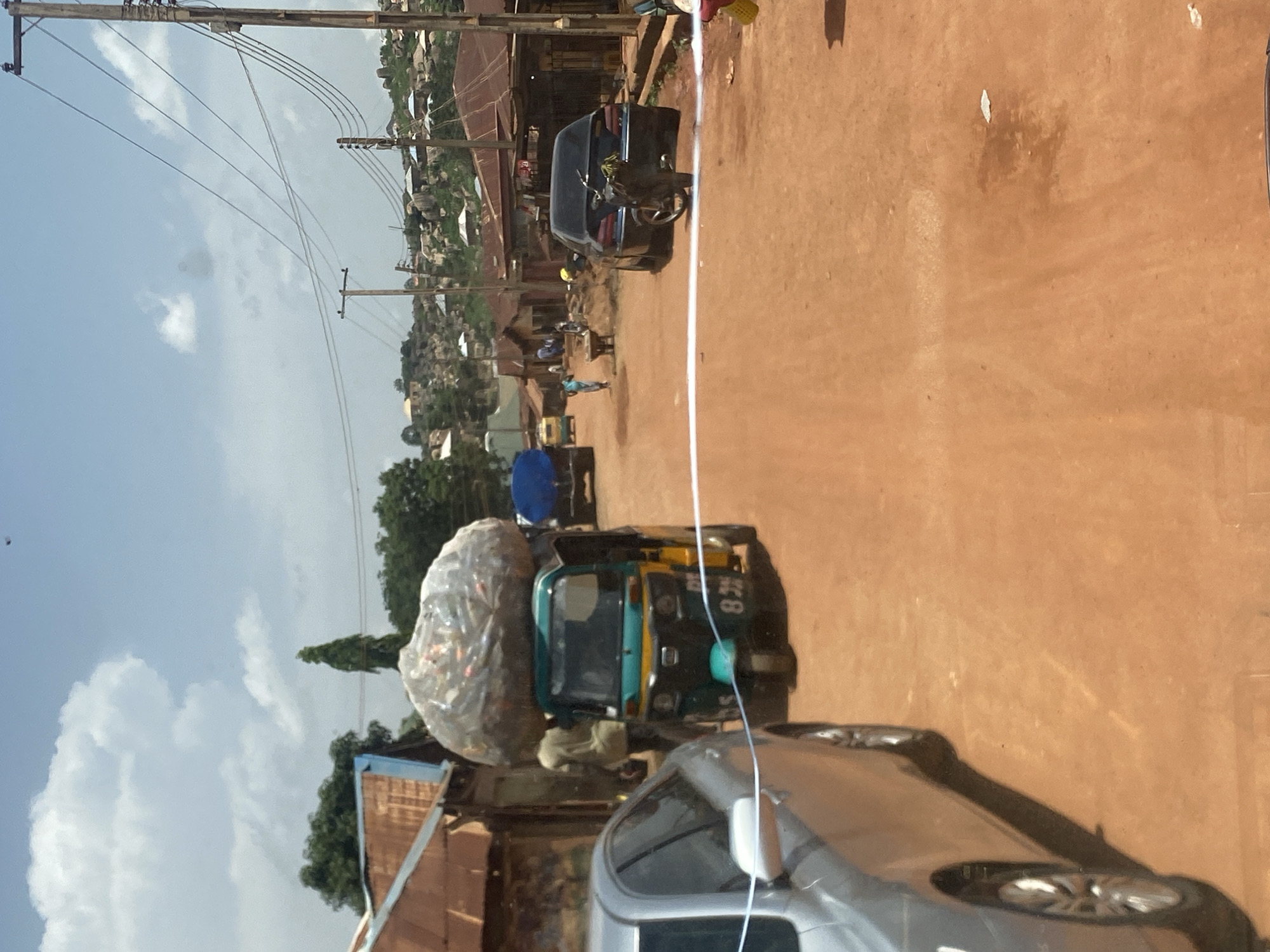 This is a street-level, eye-level photograph taken outdoors in the city of Jos, Nigeria, on a cloudy day. The scene depicts a dusty, unpaved road lined with buildings, suggesting a semi-urban or rural environment. In the foreground, a silver sedan is parked on the left side of the road. A white ribbon or tape stretches across the road, possibly indicating a barrier or event.
Dominating the mid-ground is a green and yellow auto-rickshaw, also known as a "keke," overloaded with a large, clear plastic-wrapped bundle on its roof, likely containing goods. A person, partially obscured, stands behind the auto-rickshaw. Further down the road, a blue truck is visible. Several other vehicles, including a dark-colored car and a motorcycle, are parked or moving. People are scattered throughout the scene, some walking and others engaged in activities near the buildings. The architecture of the buildings features corrugated iron roofs, typical of many structures in the region. In the background, the landscape rises into a hilly terrain dotted with more houses and vegetation. Power lines and poles are also present, indicating the presence of electricity infrastructure. The overall impression is one of daily life unfolding in a bustling, dusty environment.