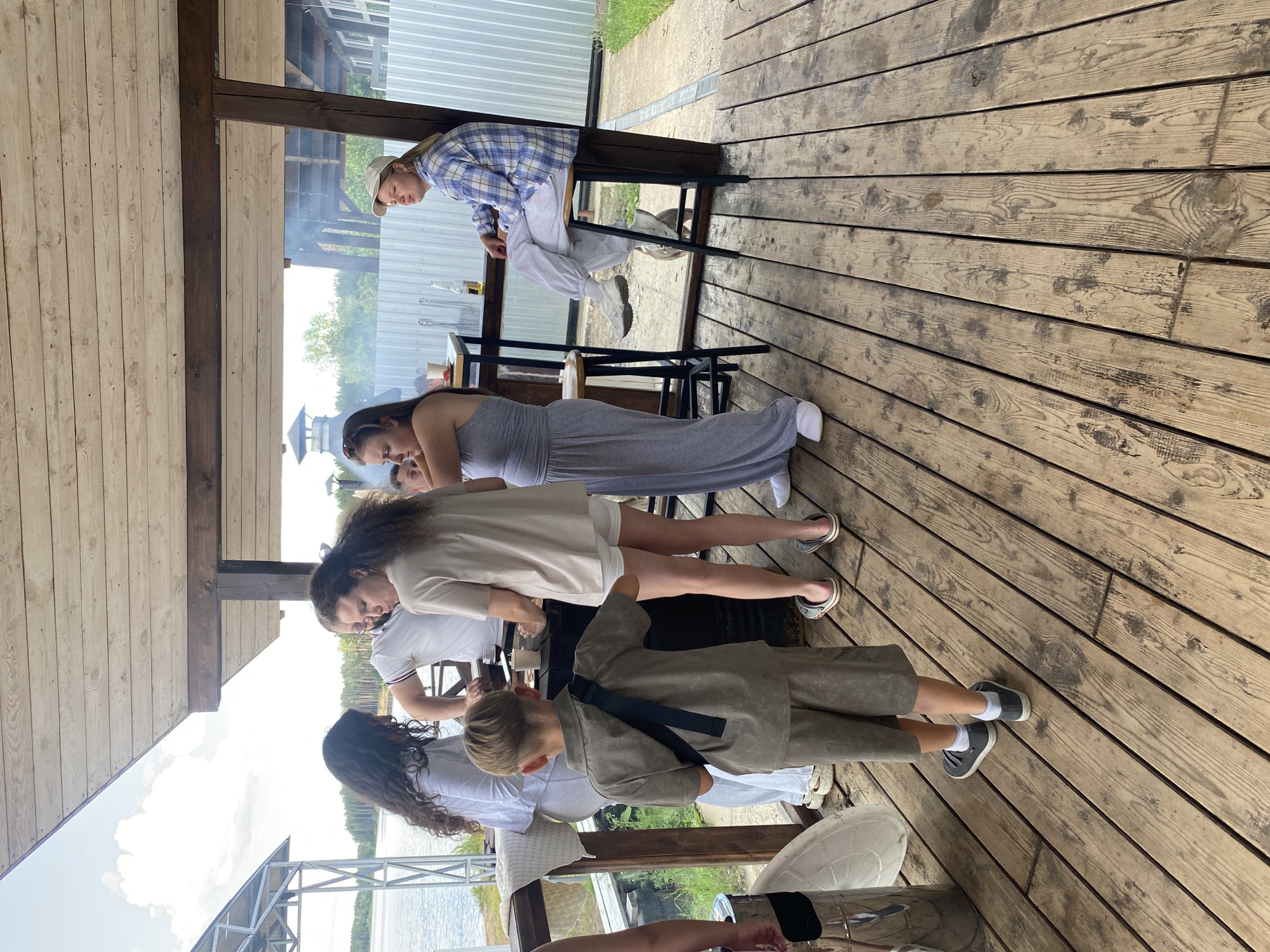 The image shows a group of people on a wooden deck, possibly at a lakeside resort, given the water and trees visible in the background. The sun is shining, suggesting it's daytime, and the weather appears pleasant. Several individuals are standing and interacting, with one person sitting on a stool, possibly engaged in a casual gathering or activity. The overall mood seems relaxed and social.
The subjects include at least five individuals, with varying ages and genders. One person, wearing a blue and white checkered shirt and grey sweatpants, is sitting on a stool. Another person in a long grey dress stands nearby. Further down the deck, a young boy in a grey outfit is interacting with others. The scene is set on a wooden deck, with a rustic, outdoor feel. In the background, there's a glimpse of water, trees, and a metal structure, possibly a pier or a boat dock.
The activity appears to be a social gathering or a casual outdoor event. There are no prominent objects that suggest a specific activity like cooking or a party.
Notable details include the rustic wooden flooring and the natural setting. The people are casually dressed, indicating a relaxed atmosphere. The lighting suggests it's daytime, and the clear sky implies good weather.
There is no visible text in the image.