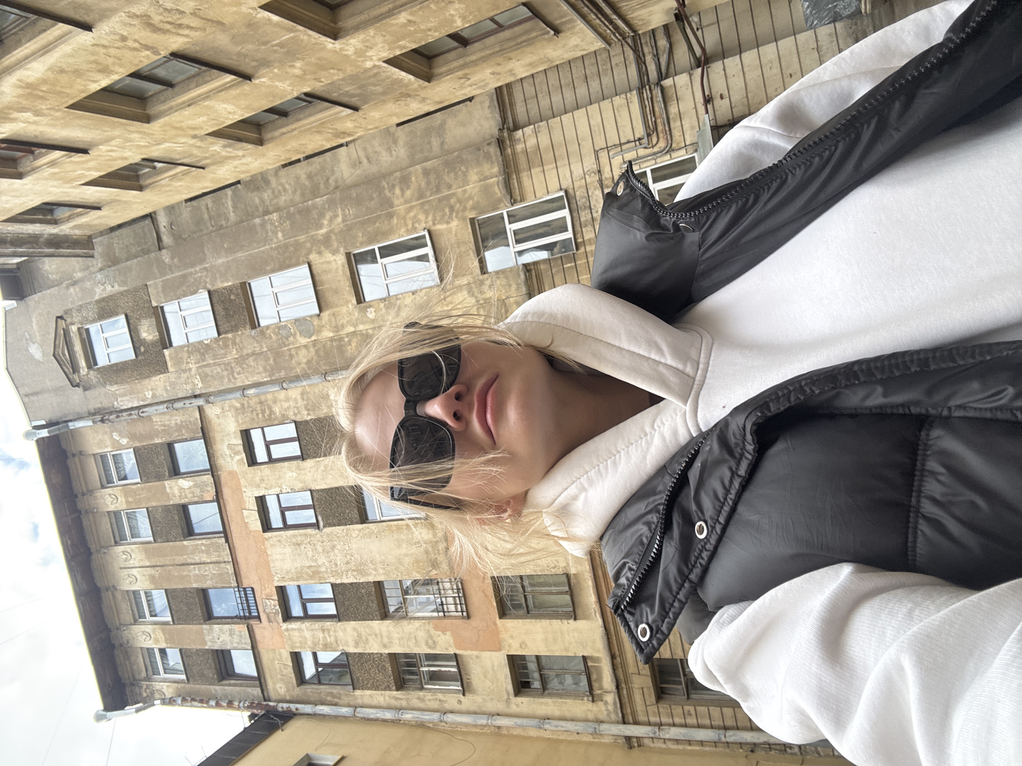 The image presents a selfie of a young blonde woman against the backdrop of an old urban building. The image is oriented with the building appearing horizontal, but when mentally rotated, it shows a multi-story facade towering behind the subject.
The central subject is a woman with blonde hair, fair skin, and full lips, wearing dark, rectangular sunglasses that cover her eyes. She is dressed in a white hooded sweatshirt layered under a black sleeveless puffer vest. The zipper and some metallic accents on the vest are visible. Her expression is neutral, possibly casual, enhanced by the sunglasses. She appears to be holding the camera herself, looking directly into the lens from a slight upward angle, consistent with taking a selfie.
The background features a densely built, aged building, characteristic of European urban architecture. The facade is a light, somewhat faded beige or ochre color, showing signs of wear and historical weathering. Numerous rectangular windows with distinct dark frames are distributed across its multiple floors. Some windows reflect the sky, while others reveal interior darkness. Architectural details include horizontal banding between floors and visible conduits or drainpipes running down parts of the building. The sky, visible in the upper left (when correctly oriented), appears bright but possibly overcast, suggesting daytime.
The location context specifies Saint Petersburg, Russia, and the architectural style of the building with its weathered facade and dense window arrangement aligns with typical older residential or commercial buildings found in that city. There is no visible text in the image.