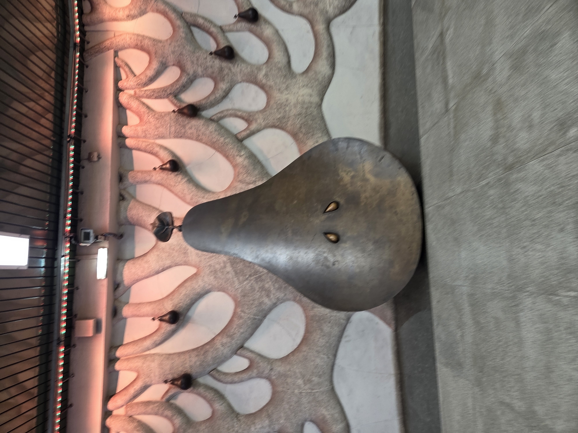 An indoor scene in an unknown location features a large, dark, pear-shaped metallic sculpture centered on a light gray panel floor. The sculpture stands upright, displaying two small, shiny, tear-drop shaped elements embedded on its upper front surface, and a small, dark, leafy detail at its apex. Behind the sculpture, a textured wall relief extends across the frame, composed of interconnected, organic, tree-like forms in a light gray hue against a lighter background. Multiple smaller, dark, tear-drop shaped objects hang from various points within this wall relief. The upper section of the wall is bathed in an orange-red glow, with a horizontal strip of alternating red and green decorative lights positioned above the relief. The ceiling above consists of parallel dark slats and includes rectangular fluorescent light fixtures. No people are present.