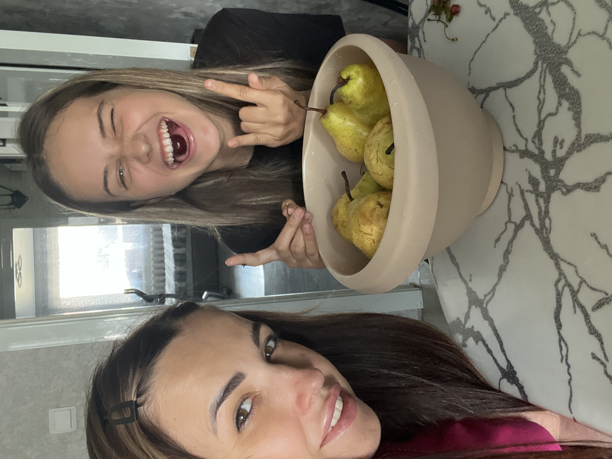 In an indoor environment, two young women are captured in a selfie, positioned behind a bowl filled with pears. The woman on the left, with long brown hair, is looking towards the camera with a gentle smile. The woman on the right, also with long blonde hair, is laughing expressively, winking, and making the "rock on" hand gesture. She appears to have a cherry or similar small red fruit in her mouth. The bowl of pears sits on a marble-patterned surface, suggesting a kitchen or dining area. The background shows a doorway leading to another room with a window and furniture, hinting at a domestic setting. The lighting suggests it is daytime.