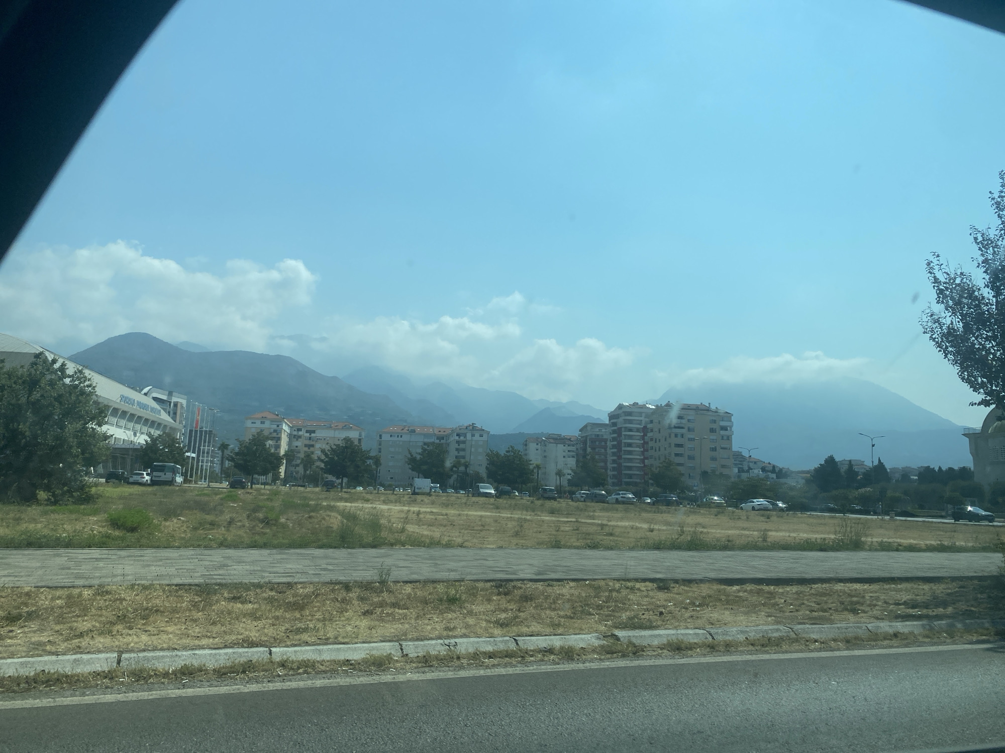 The image, taken from a moving vehicle, depicts a city skyline against a backdrop of hazy mountains. In the foreground, a dry, grassy median strip separates the road from a paved walkway. Beyond the walkway, several buildings of varying heights and architectural styles are visible, interspersed with trees and parked cars. A large, modern white building with a curved roof, possibly a sports arena or convention center, is prominent on the left side of the frame. The sky is a pale blue with scattered clouds. The general impression is an urban environment in a mountainous region, likely during the daytime. The location appears to be the city of Bar, Montenegro, given the characteristic architecture and mountainous terrain often seen in the area. There are no visible people or specific actions being performed.