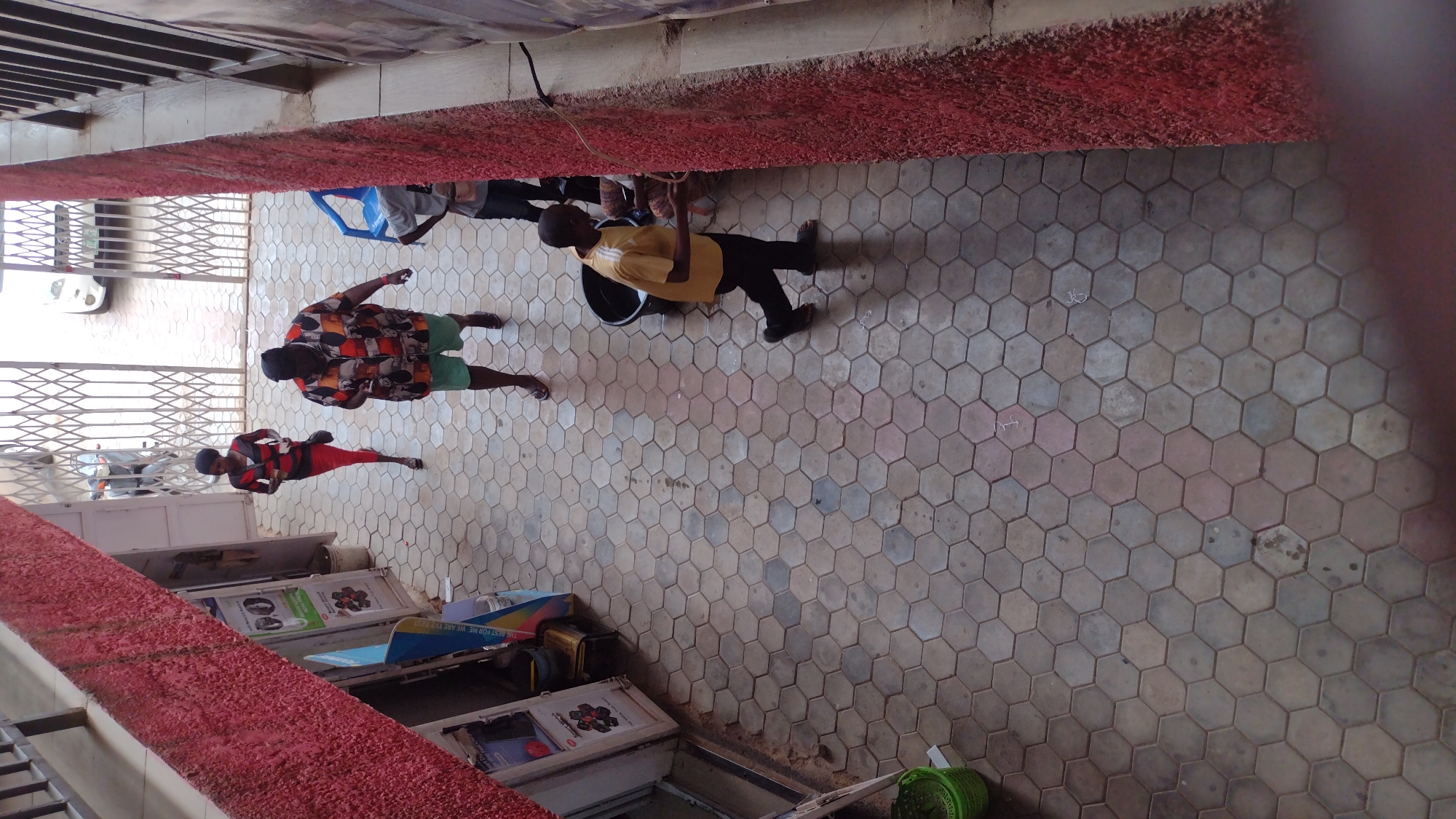 The image captures an outdoor scene in an urban environment, specifically in Unknown, Unknown. The ground is covered in hexagonal paving stones of varying shades, creating a textured surface. A red-painted ledge runs along the top and left side of the frame, indicating the presence of a building or structure.
Several people are visible in the scene. One person is wearing a yellow shirt and dark pants, carrying what appears to be a black bucket. Another individual is dressed in a colorful patterned shirt and green shorts. A third person is wearing a red and black outfit. All three are standing or walking on the paved area.
Along the left side of the frame, several display cases are visible, some containing advertisements or product information. Behind them, there is a metal gate. In the background, glimpses of a white car are visible. A green plastic basket is on the right-hand side of the shot.