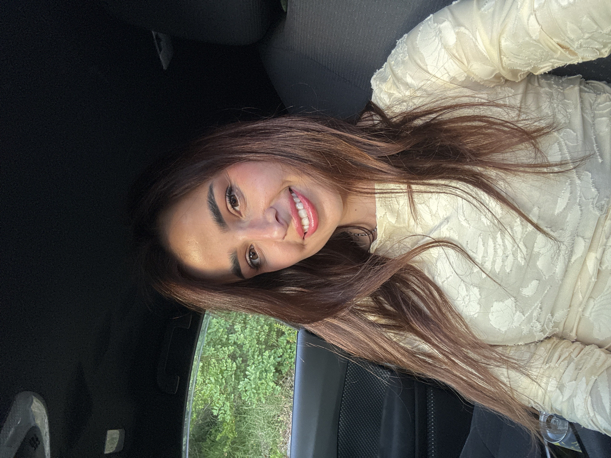 The image displays a close-up, angled selfie of a young woman smiling brightly from inside a car. She has long, flowing dark brown hair with reddish highlights that falls over her shoulders. Her face is well-lit, revealing prominent makeup including defined eyebrows, winged eyeliner, and glossy pink lipstick, accompanied by rosy blush on her cheeks. She is smiling broadly, showing her white teeth.
The woman is wearing a light-colored top, possibly cream or off-white, which features a textured, embossed, or woven floral pattern across the fabric. The sleeves appear to be gathered or ruched.
The setting places her inside a vehicle, with parts of a dark car seat and the dark headliner visible in the background. Through the car window, seen on the left side of the frame, there is lush, vibrant green foliage, indicating an outdoor, natural environment during the daytime. The sunlight illuminates her face, creating a cheerful and friendly expression. No discernible text is visible in the image.