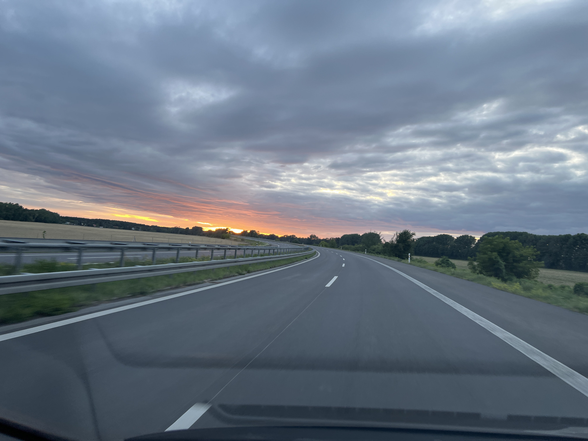 The image depicts a highway scene at sunset in the vicinity of Trebbin, Germany. A dark asphalt road curves to the right, with white lane markers. A metal guardrail runs along the left side of the road, separating it from a grassy verge and a field beyond. The sky is filled with dramatic clouds, tinged with orange and yellow hues from the setting sun on the left side of the frame. Trees line the horizon on both sides of the road. The perspective suggests the image was taken from a vehicle driving on the highway.