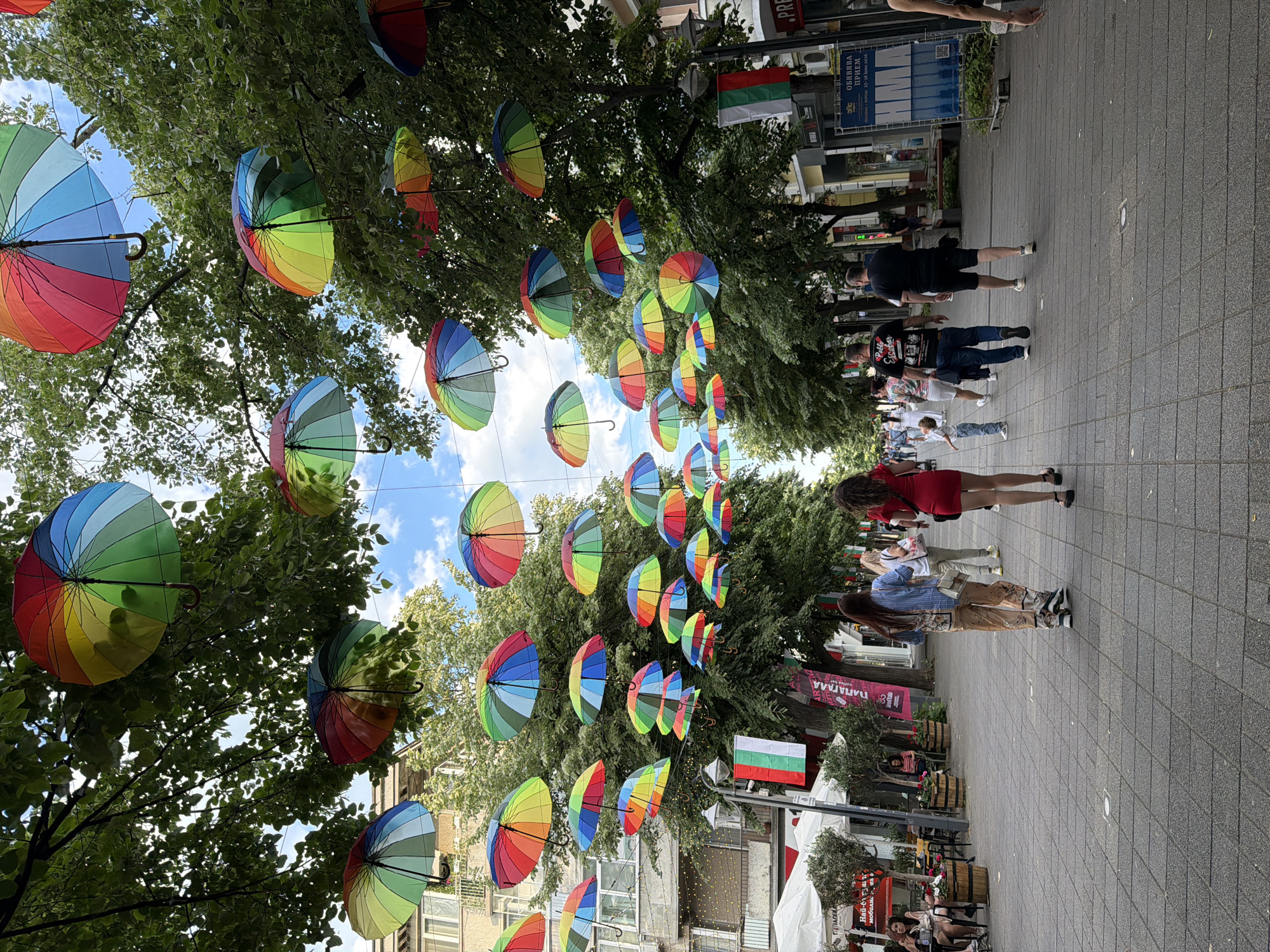 The image depicts a pedestrian street in Burgas, Bulgaria, adorned with rows of colorful, open umbrellas suspended above the walkway. People are walking along the street, some wearing masks. The street is lined with shops and restaurants, and the Bulgarian flag is visible. Signage is present with text in Bulgarian. The surface of the pedestrian walkway is paved with gray, rectangular stones.