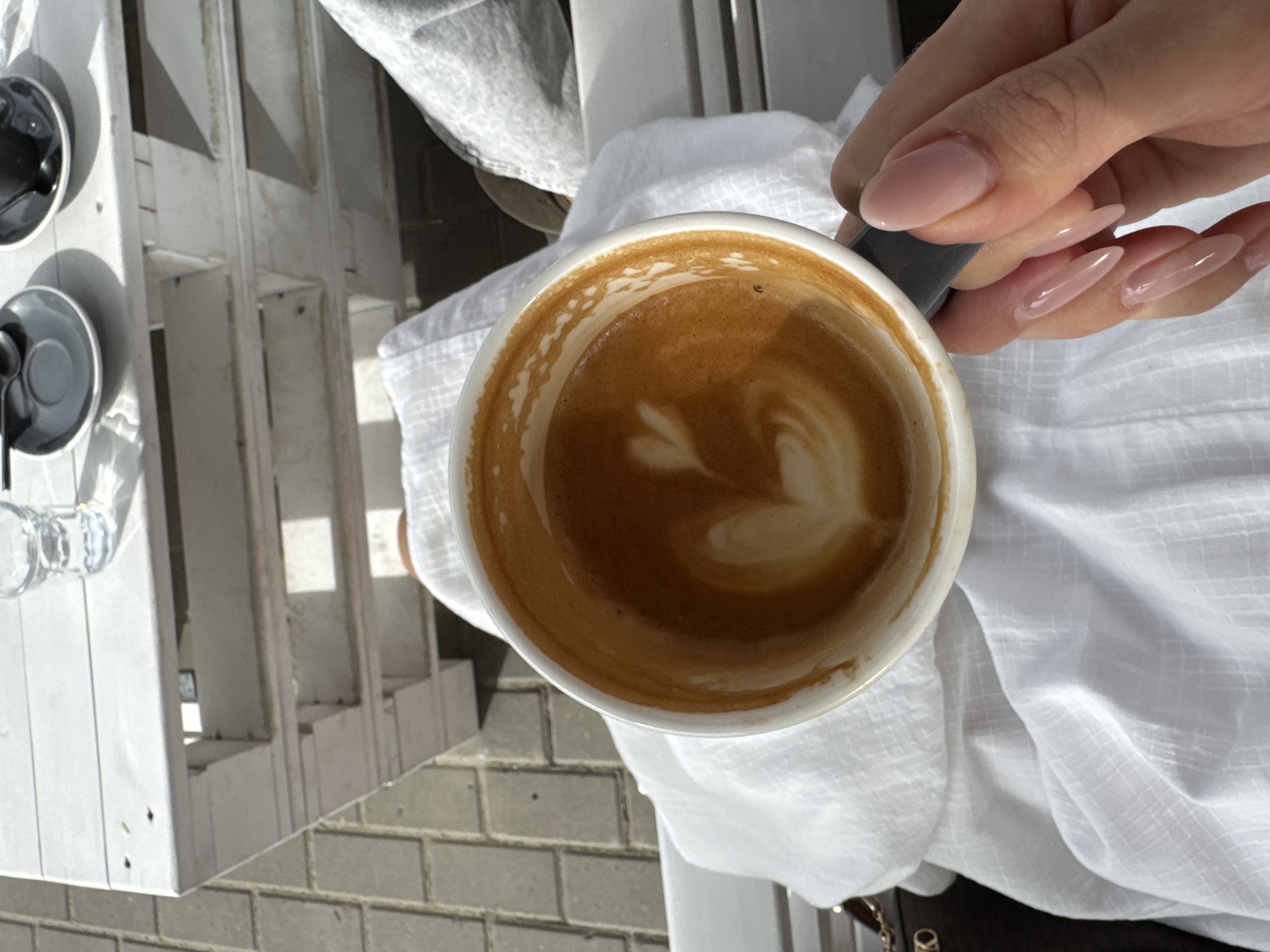 A close-up, top-down shot shows a hand holding a cup of coffee with latte art. The coffee cup is white and has a dark grey handle, and the latte art appears to be a heart shape. The hand holding the cup has long, manicured nails painted with a light pink color. The cup is placed on a white linen fabric. In the background, there is a table with white wooden slats and some other cups and saucers, and a drinking glass. The floor appears to be paved with grey bricks. The location is outside on a sunny day.