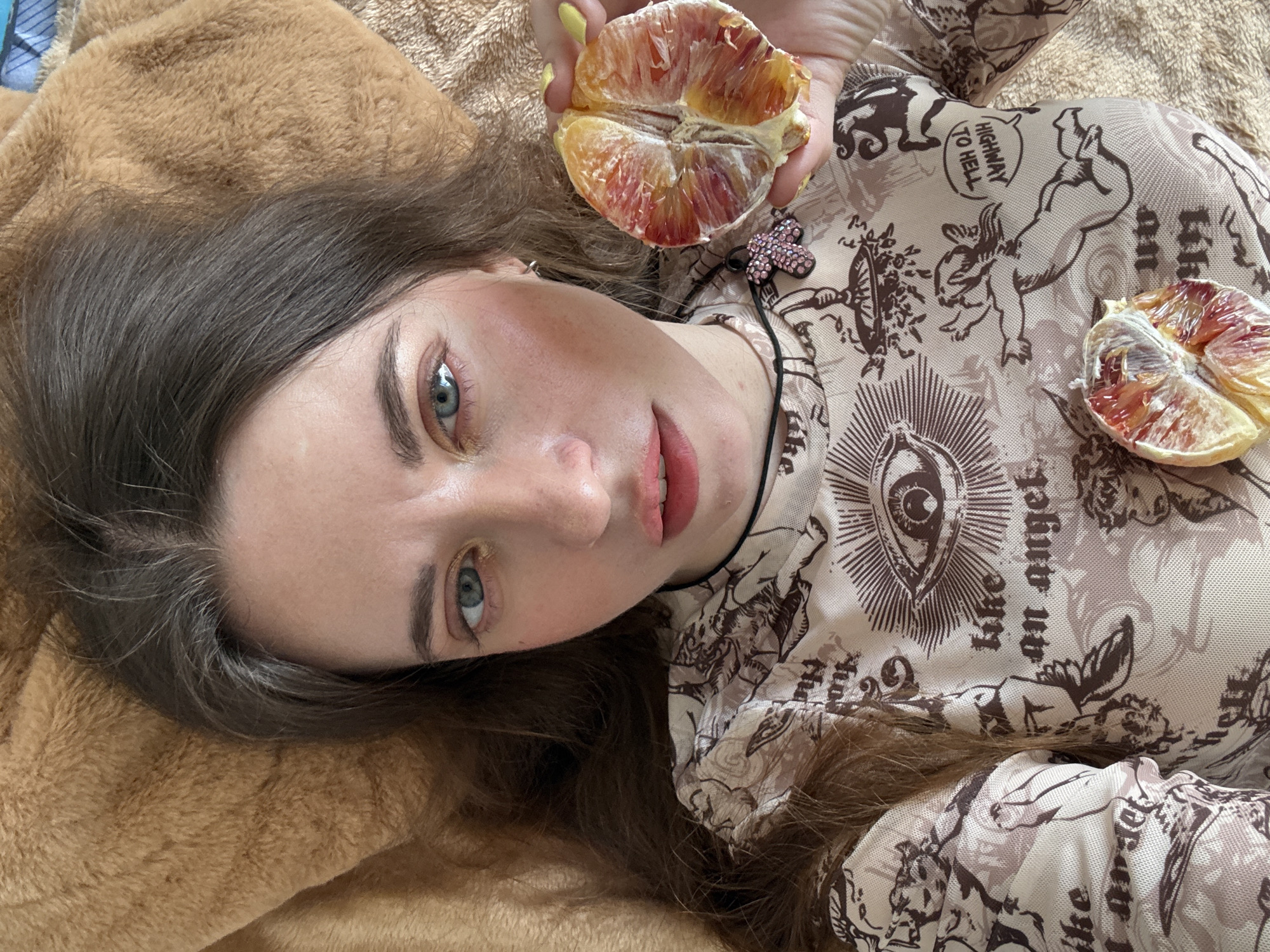 A young woman is lying on a brown, textured surface, possibly a bed or blanket. She has fair skin, light blue eyes, and brown hair. She is wearing a patterned top with motifs of angels, eyes, and the phrase "Highway to Hell." She is holding a halved citrus fruit, possibly a blood orange, in her left hand. A second halved blood orange is on the right side of the frame near her torso. The background is blurred. No information to indicate the location as Moscow, Russia.