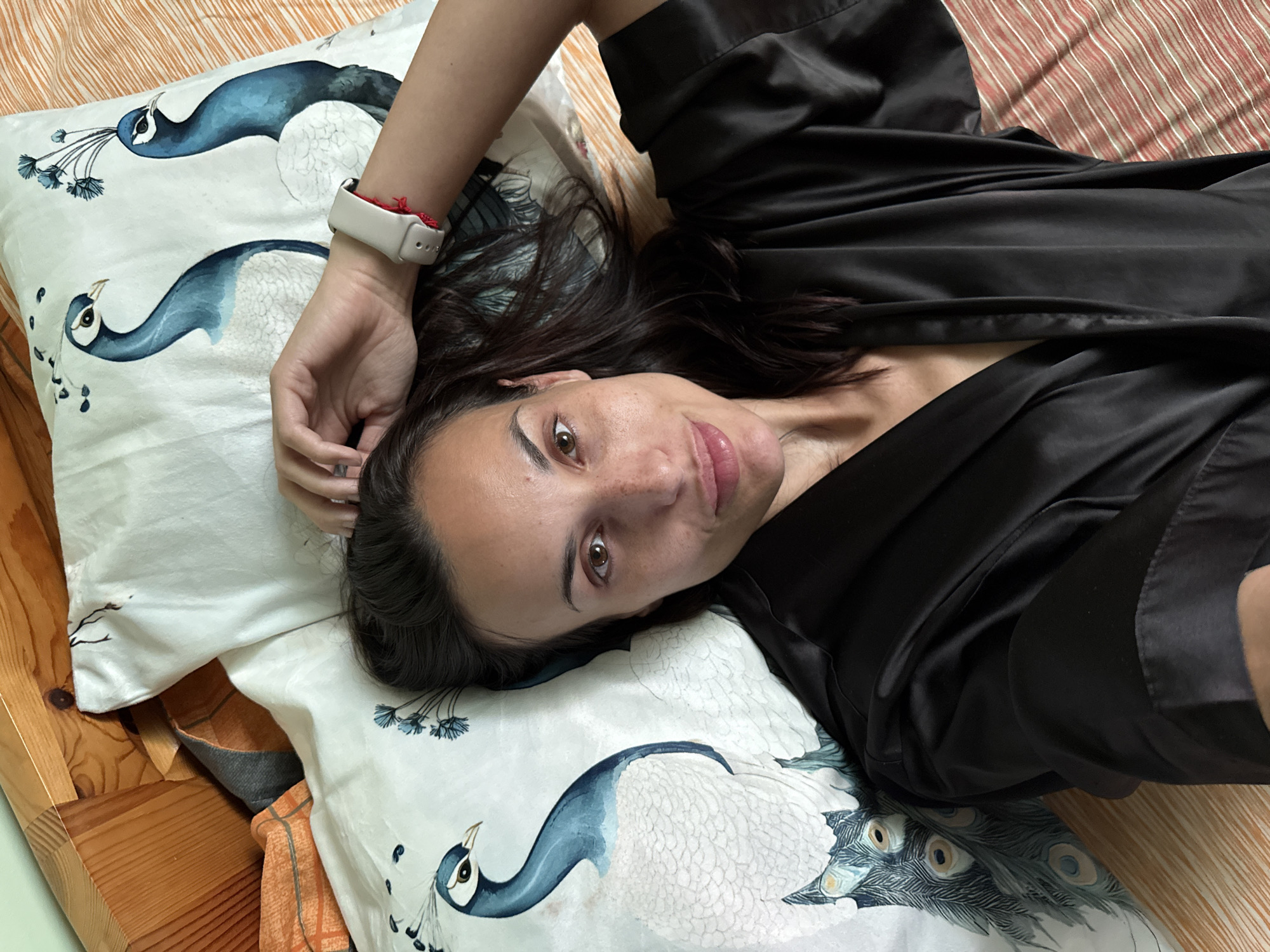 A woman is lying on a bed, wearing a black satin robe and a smartwatch. She is looking at the camera and has her hand raised to her head. The bed has pillows with a peacock design, and the sheets are striped. The background shows a wooden headboard and other bedding. The scene appears to be indoors, likely a bedroom.
