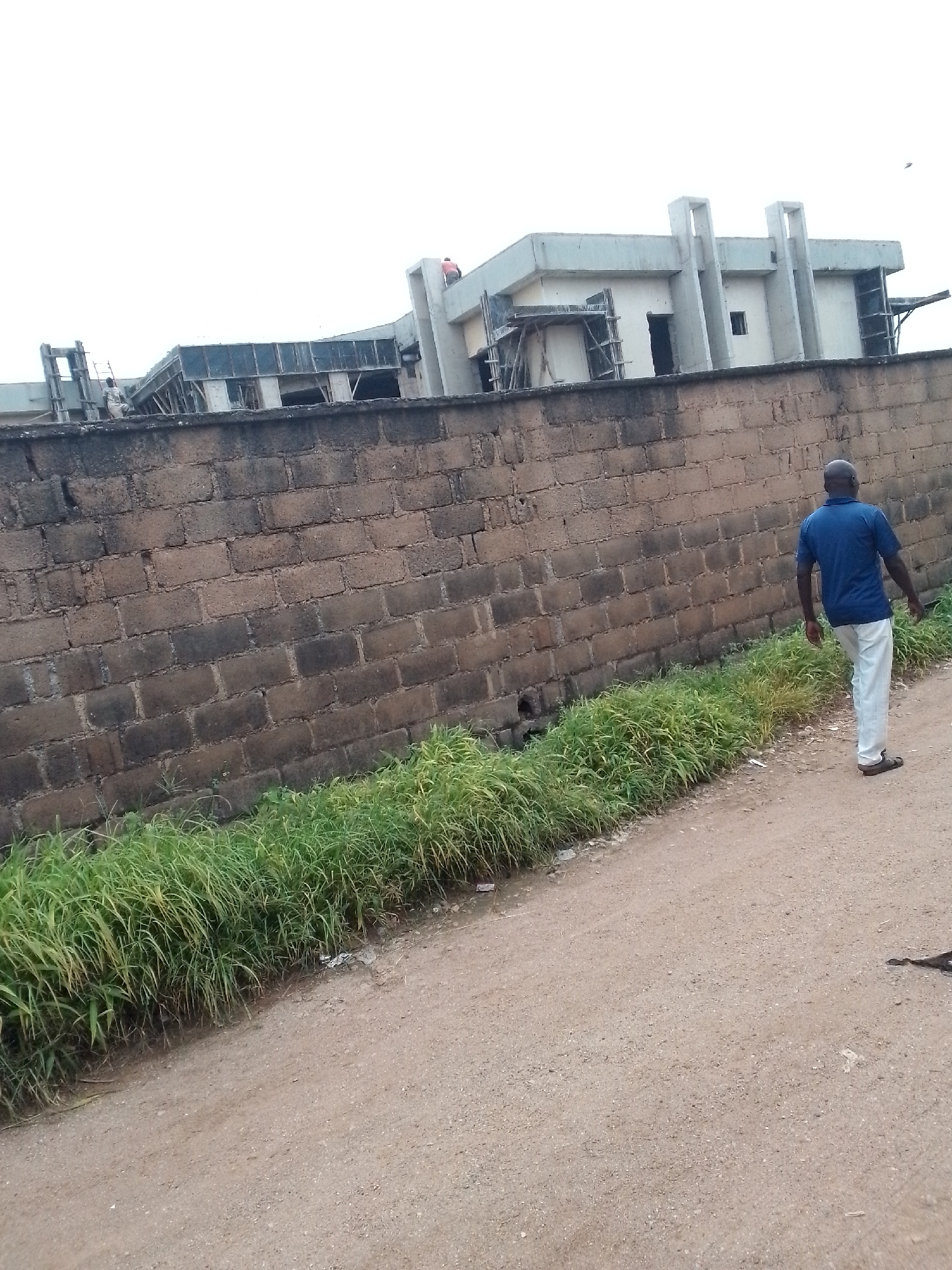 A bald Black man wearing a blue short-sleeved polo shirt and off-white trousers walks away from the camera on a dirt road. He is on the right side of the frame, with his back to the viewer. He is wearing dark sandals. To his left is a low wall constructed of rough, dark bricks. Beyond the wall is a building under construction, featuring concrete structures and scaffolding. A narrow strip of tall, green grass runs along the base of the brick wall, bordering the dirt road. The sky is overcast and white. The scene appears to be in an undeveloped or construction area in Jos, Nigeria.
