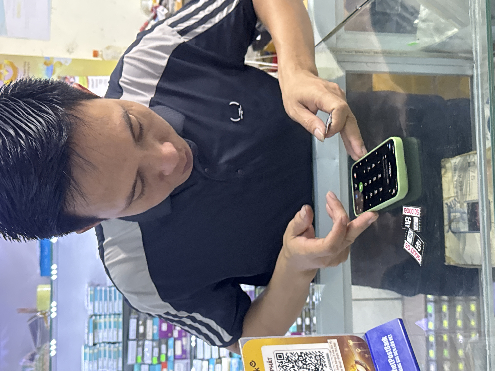 A man in a dark shirt with white stripes on the shoulders is standing behind a glass counter. He is focused on a smartphone with a green case, holding it with both hands. The screen of the phone is visible, displaying numbers and icons. Stickers are stuck to the glass counter. Below the phone, there is a yellow promotional item. Behind the man, there are shelves stocked with various merchandise, suggesting a store setting. The OCR text suggests that this image was taken at Du Già Commune, Vietnam.