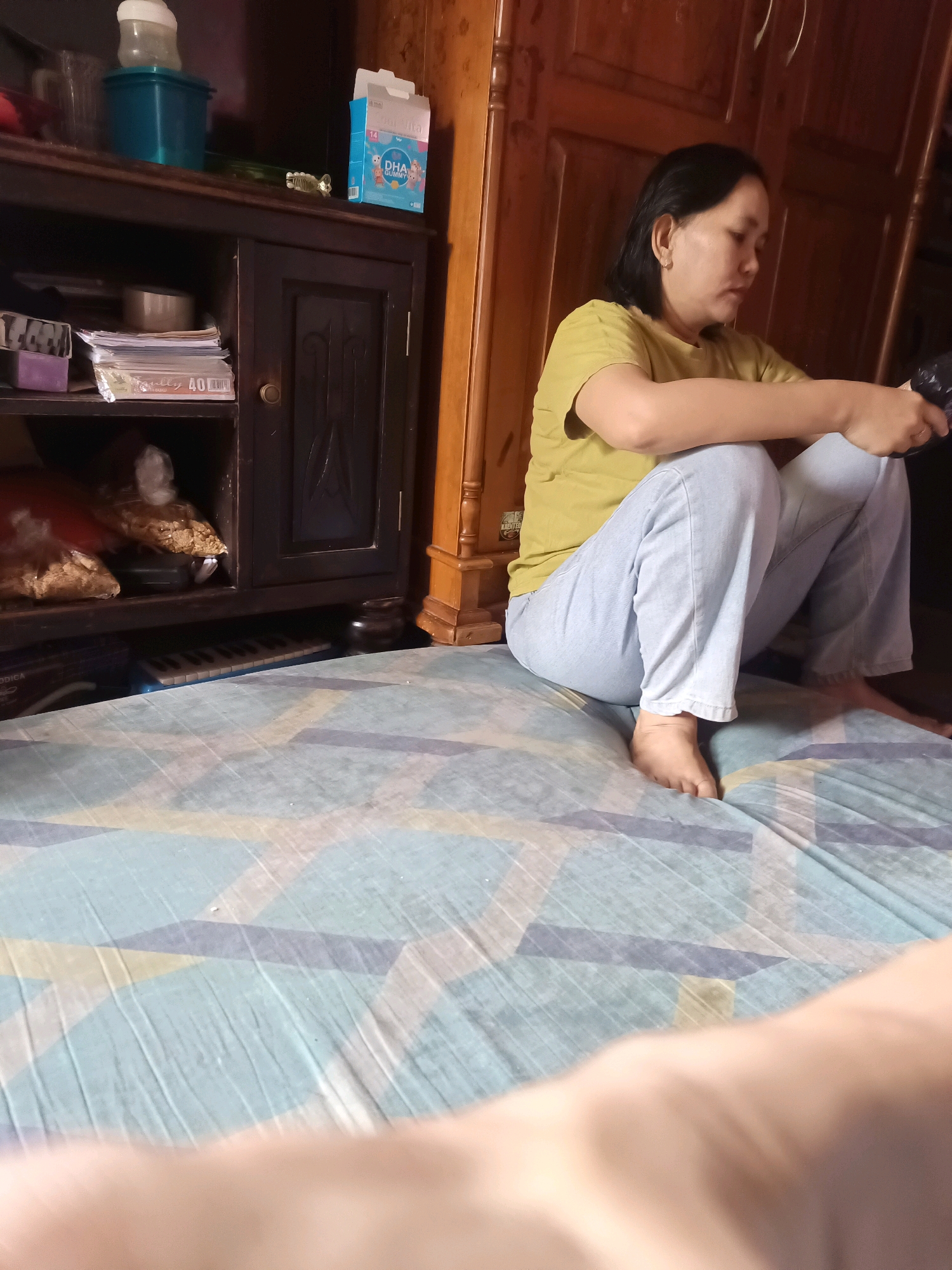An Indonesian woman is seated cross-legged on a blue and yellow patterned mattress, facing to her right. She is wearing a yellow t-shirt and light blue jeans. Her hair is dark and short, and she has a small earring in her left ear. She is holding a black object in her hands, possibly a phone or a remote control. The background consists of a dark wooden cabinet with shelves containing various items, including boxes of DHA Gummy, a baby bottle, and stacks of papers. To the right of the cabinet, a wooden pillar is visible. The floor beneath the mattress is not visible. The scene appears to be indoors, in a residential setting.