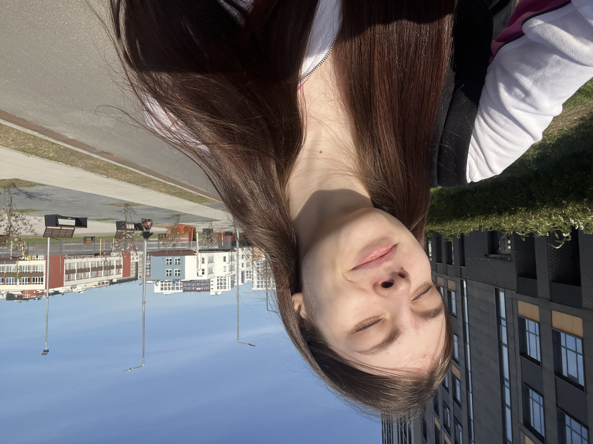 A young woman with long brown hair is outdoors on a sunny day in Saint Petersburg, Russia. She wears a white top under a black jacket. The location appears to be a modern urban area with multi-story residential buildings, streetlights, and benches. The sky is blue.