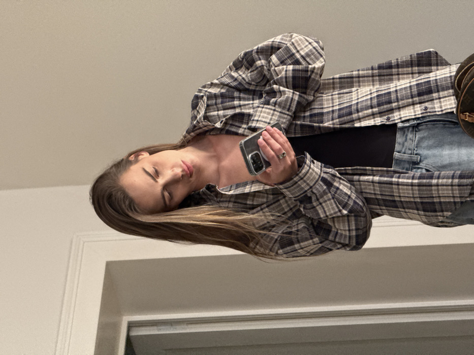 A female individual with long brown hair is depicted indoors, approximately from the waist up, engaged with a smartphone. She wears a blue, white, and brown plaid button-up shirt, unbuttoned, over a black top, and light blue denim jeans. A ring is present on her left ring finger. Both hands hold a dark-colored smartphone, featuring a multi-lens camera array on its rear, encased in a clear protective cover, with her gaze directed downwards towards the screen. The background consists of plain, light-colored walls. A white door frame or architectural trim is visible on the left side of the frame. A portion of a brown patterned bag is visible at the lower right. There are no discernible elements within the image to indicate the specific location is the city of Venice, Italy; the environment appears to be a generic interior space.