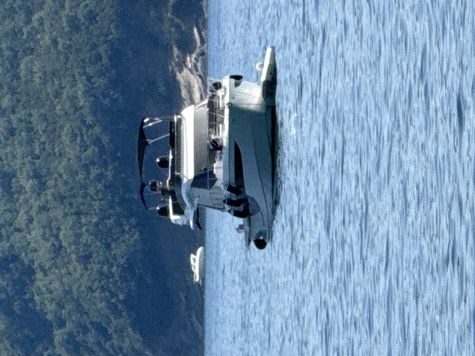A modern, multi-level white yacht with a black canopy is docked on calm, blue water, with a lush green forested hill in the background. Another smaller white boat is visible in the distance to the left. The image is taken from a medium distance, showing the yacht from the front-port side. The water is rippled, indicating a gentle current. The overall scene is serene and depicts a leisurely day on the water.