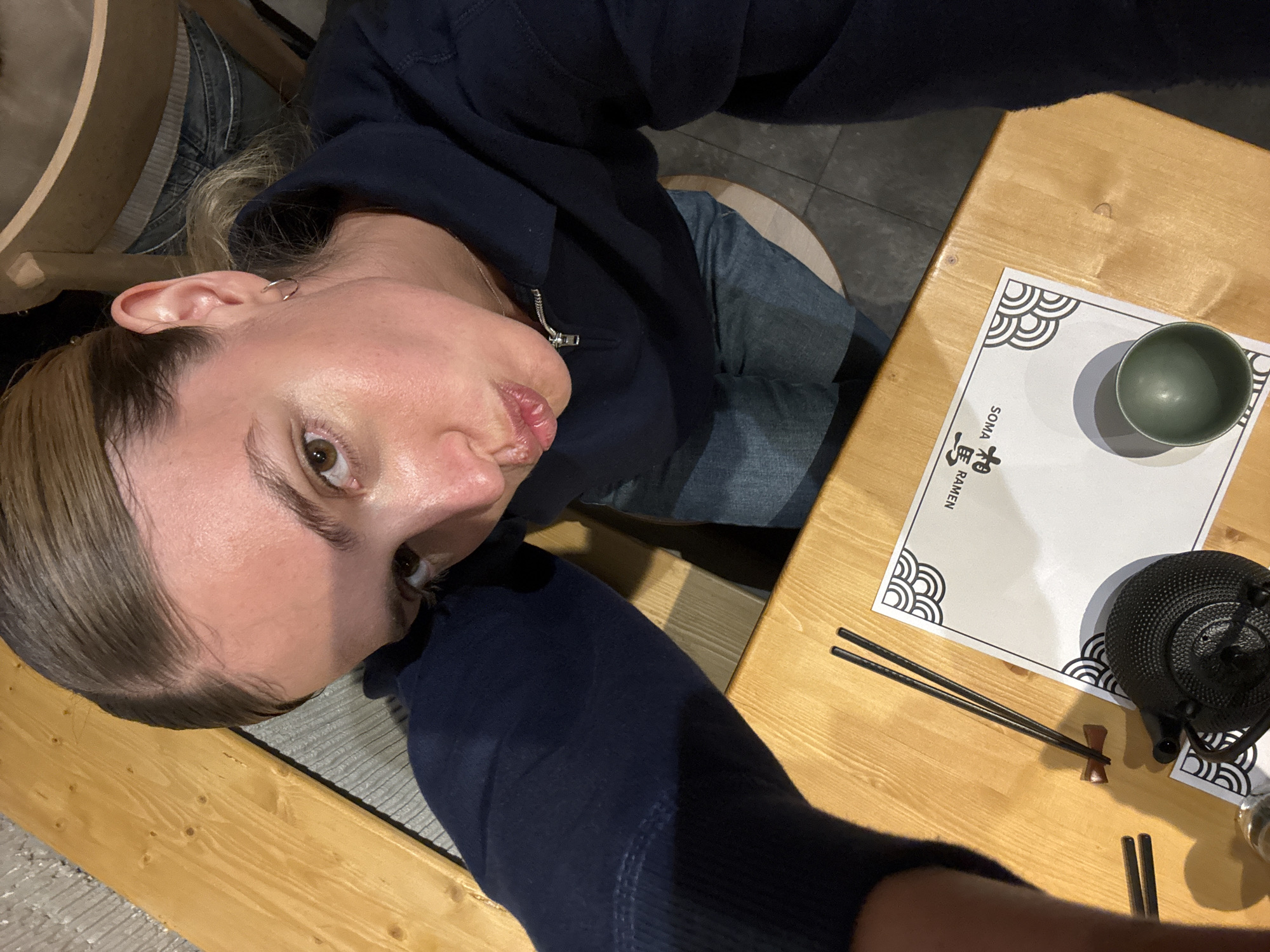 A selfie taken by a woman in a navy blue hooded jacket while at a restaurant. She is seated at a light wooden table. On the table is a place mat with black and white decorative designs and the name "SOMA Ramen." There is a green bowl, a black cast iron teapot, and a pair of chopsticks. The woman has light brown hair pulled back, fair skin, and light brown eyes. She has a small silver hoop earring in her left ear. The photograph appears to be taken from a slightly upward angle. SOMA Ramen is a restaurant in Tbilisi, Georgia.