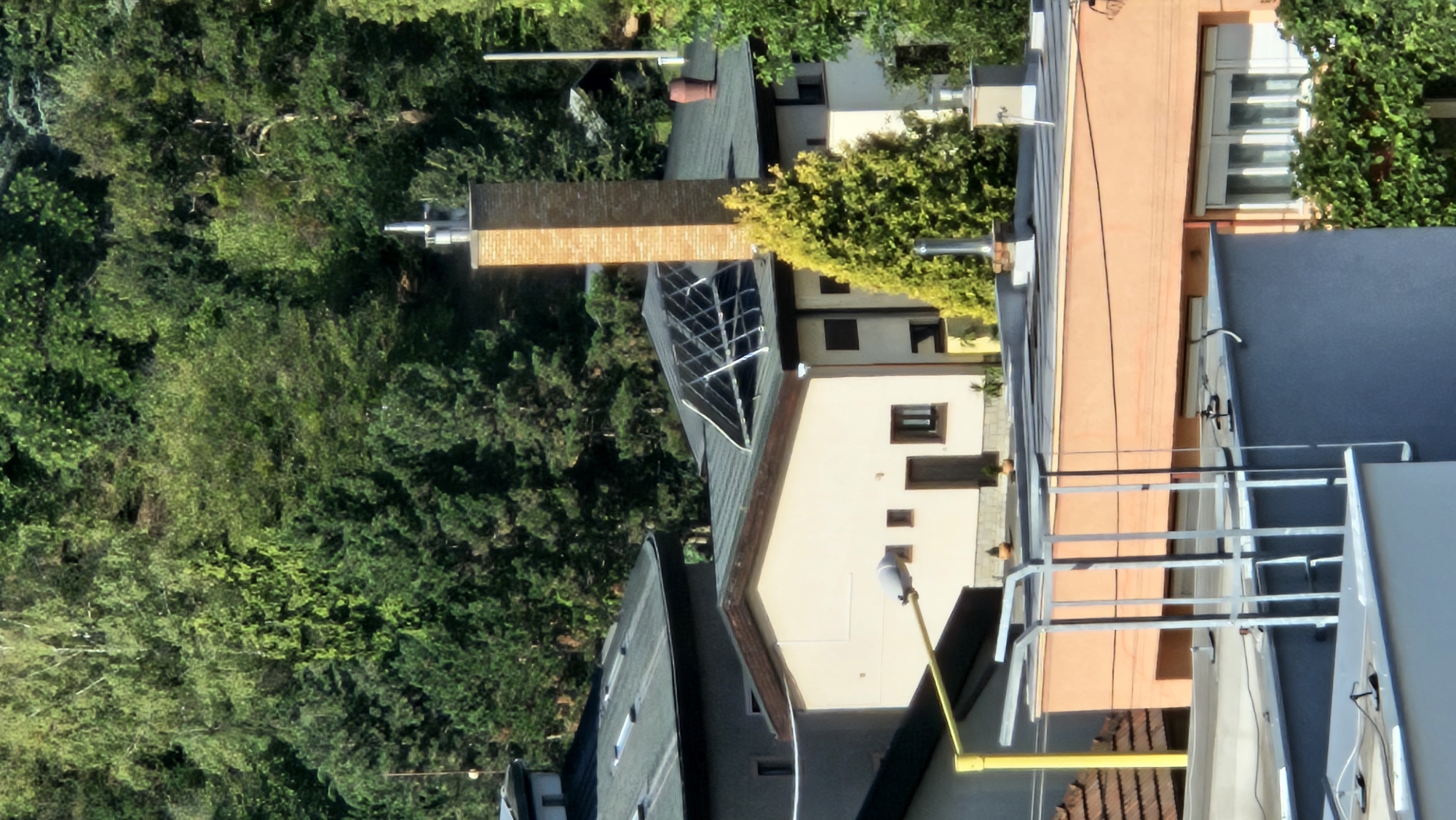 The image shows a residential area with several houses and buildings nestled into a hillside covered in dense green trees. The buildings have varied roof lines and architectural styles. One building in the mid-ground features a prominent brick chimney with a metallic cap. Another building has a dark sloped roof with what appears to be a solar panel array. In the foreground, there are sections of blue and orange-colored building facades, with some metal structures and windows visible. The overall scene suggests a suburban or semi-rural environment on a sunny day. Based on the context of the city of Prešov, Slovakia, this image likely depicts a residential neighborhood on the outskirts or in a hilly part of the city, which is known for its proximity to the Slanské vrchy mountains.