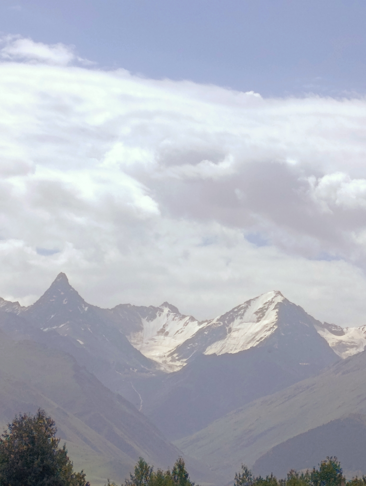 The image depicts a mountain range with snow-capped peaks under a cloudy sky. The mountains are steep and rocky, with patches of snow visible on their summits and slopes. The foreground shows trees and vegetation, suggesting a natural landscape. The scene appears to be a clear day, with the sun casting light on the mountains. The image's overall tone is serene and majestic, highlighting the grandeur of nature. The location is At-Bashy, Kyrgyzstan.