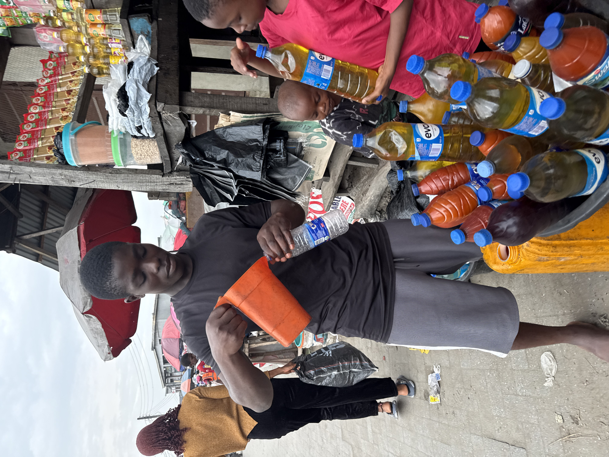 In a street market in Ado, Nigeria, a man wearing a black t-shirt and gray shorts stands barefoot, pouring water from a clear plastic bottle into an orange measuring cup. He is positioned in front of a pile of plastic bottles filled with various liquids, some labeled "eva" and "Swal." A young child in a red shirt stands near the bottles, possibly assisting. Behind the man, a woman in a gold-colored top and black pants holds a black plastic bag. Various other vendors and shoppers can be seen in the background, under umbrellas and makeshift stalls. A variety of food items, like spaghetti and cooking oil, are displayed on shelves above.