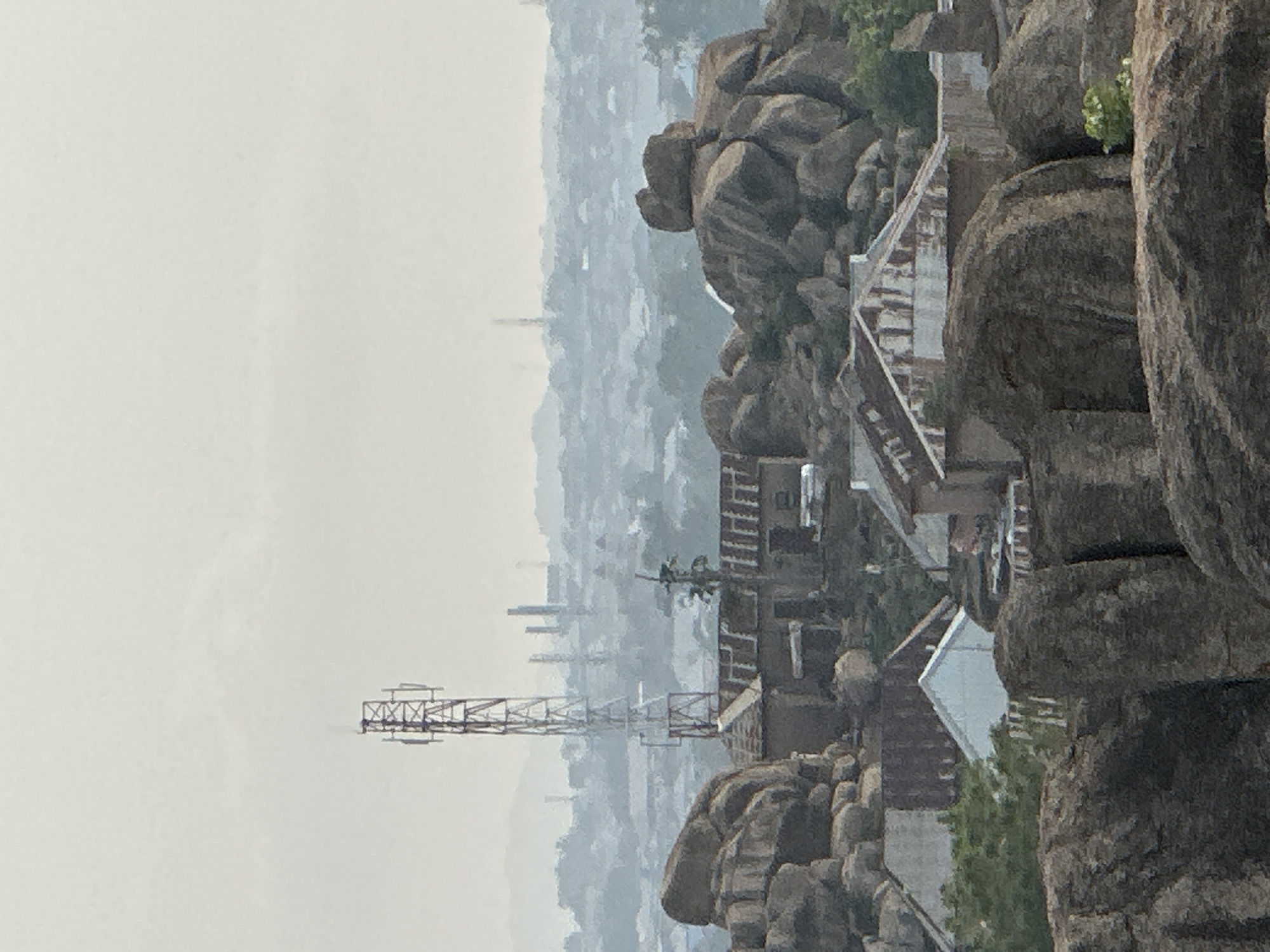 The image displays an elevated, wide-angle view of a rocky landscape overlooking a hazy city, consistent with the Jos, Nigeria context. The foreground and right side are dominated by large, dark gray-brown boulders and rock formations, with occasional patches of green vegetation sprouting from crevices.
In the mid-ground, several structures are integrated into the rocky terrain. These include buildings with light-colored walls and dark roofs, and a series of staircases or paths winding upwards among the rocks. A prominent, tall metal lattice tower, likely a cell tower or communications mast, stands on the left side of the mid-ground, extending towards the sky.
The background presents a sprawling urban area, appearing indistinct and blurry due to significant atmospheric haze or mist. The city buildings are silhouetted against a uniformly light gray or off-white sky, suggesting an overcast day. No specific human activity or people are visible within the frame. The time of day appears to be daytime, characterized by diffuse light. There is no discernible text in the image.