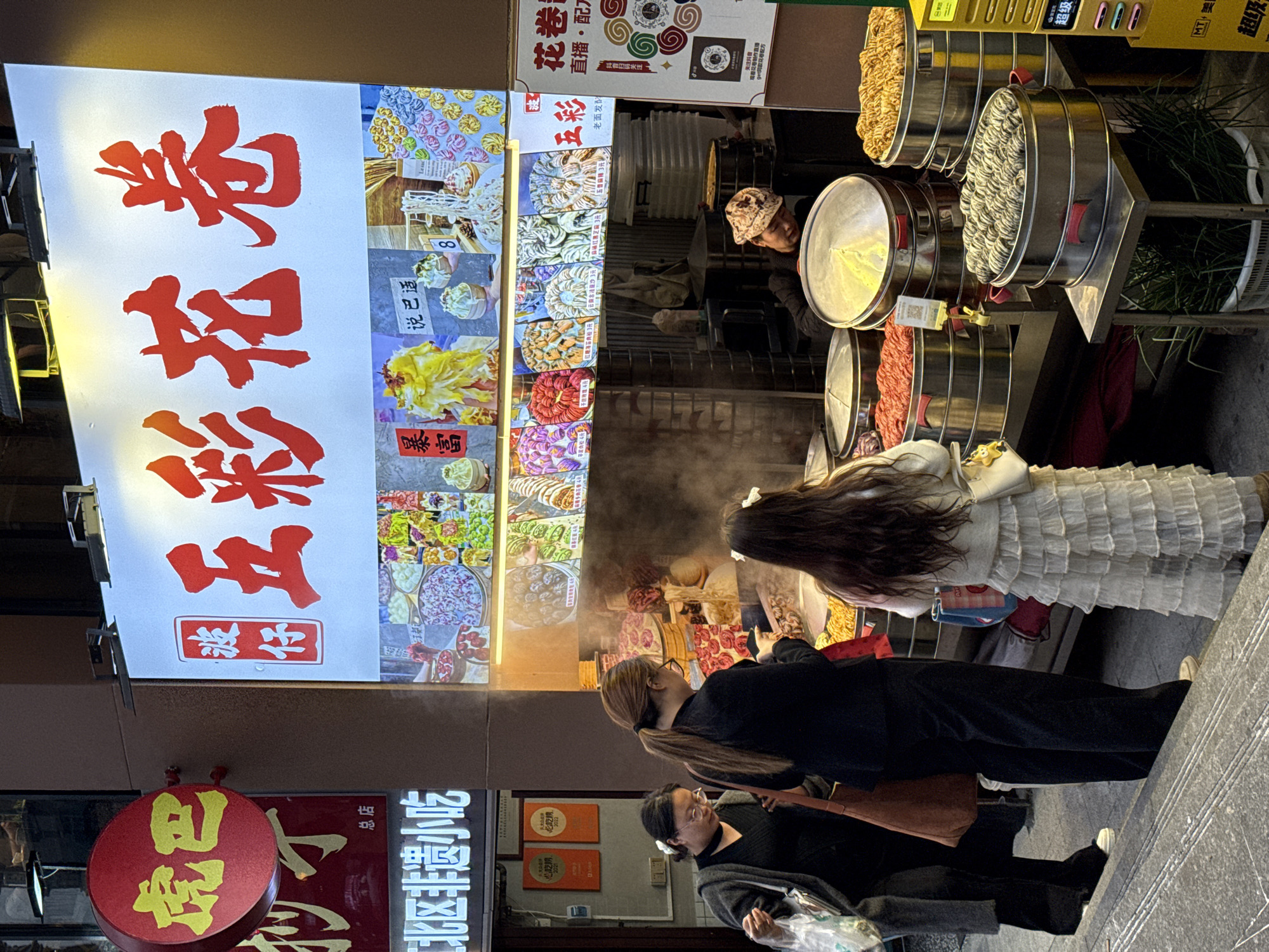 Here is a description of the image:
The image shows a street food vendor in Liangjiang, China. The storefront has a large sign with Chinese characters and pictures of the different types of dumplings they sell. There are three women standing in front of the vendor, seemingly looking at the food and about to order. Behind the vendor, there are large metal pots with different types of dumplings. The scene is filled with the steam of cooking food.