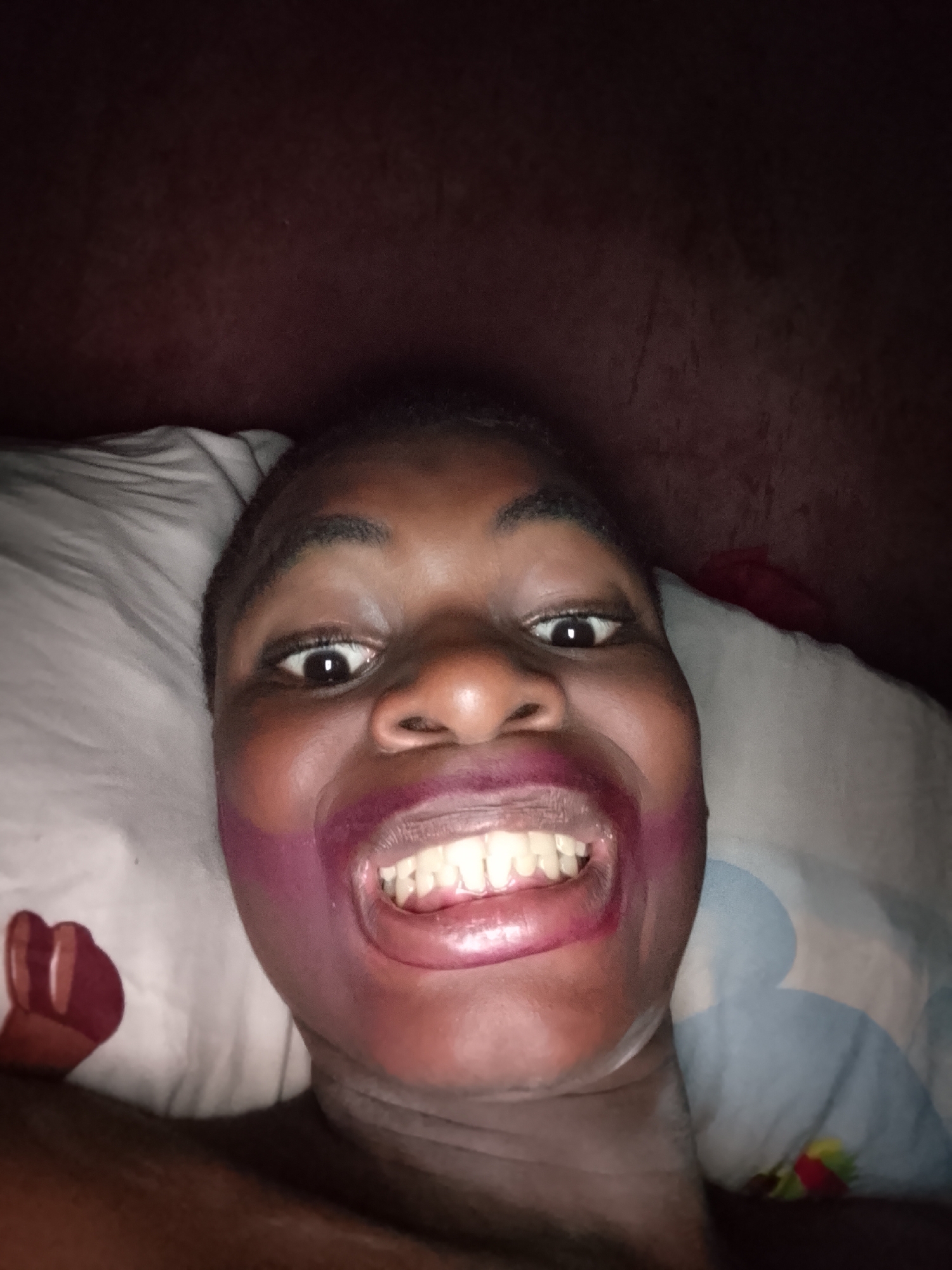 A close-up, high-angle image captures a person's face, likely a selfie, with their head resting on a pillow. The person has dark skin and short, dark hair, and is looking directly at the camera with wide, bright eyes. A broad, open-mouthed smile reveals a full set of teeth.
A prominent detail is the smudged purple or dark pink substance around the person's mouth, extending onto the cheeks, suggesting uneven application or smudging of lipstick or a similar cosmetic.
The person is lying on a light-colored pillow, which features faint patterns. On the left side of the pillow, a stylized red shape, possibly a heart or part of a larger design, is visible. On the right, a muted blue pattern with small colorful accents can be discerned. The background behind the person's head is a dark, solid surface, appearing to be a dark red or brown, possibly a headboard or wall.
The scene appears to be indoors, likely in a bedroom. The lighting is low overall, suggesting evening or night, with the person's face brightly illuminated by an artificial light source, such as a camera flash, creating strong contrasts. The person's expression conveys a sense of playfulness, surprise, or joy. No text is visible in the image.