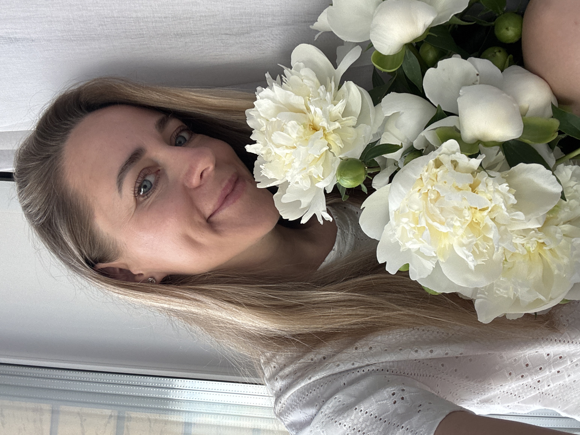 A blonde woman with blue eyes is holding a bouquet of white peonies, with a window and curtains in the background. The scene is indoors, likely a home, and the woman is looking directly at the camera with a gentle smile. The lighting is soft, suggesting natural light. There is no indication of the city of Chaikovsky, Russia.