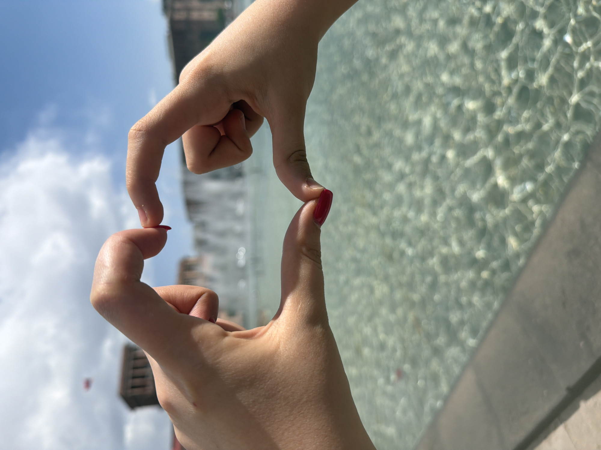 The image shows two hands forming a heart shape in front of a water feature. The fingernails are painted red. The background is blurred, but the water feature appears to be a shallow pool with textured water surface, and a building can be seen in the distance. The sky is visible above the building with some clouds. The scene is in Yerevan, Armenia.