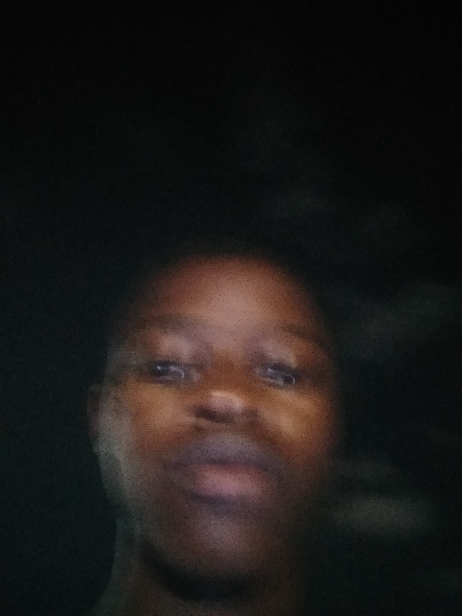 The image is a close-up of a person's face. The person has dark skin, dark eyes, and dark hair. The background is dark and blurry. The photo was taken in Shangisha, Nigeria. The face is in focus while everything else is out of focus.