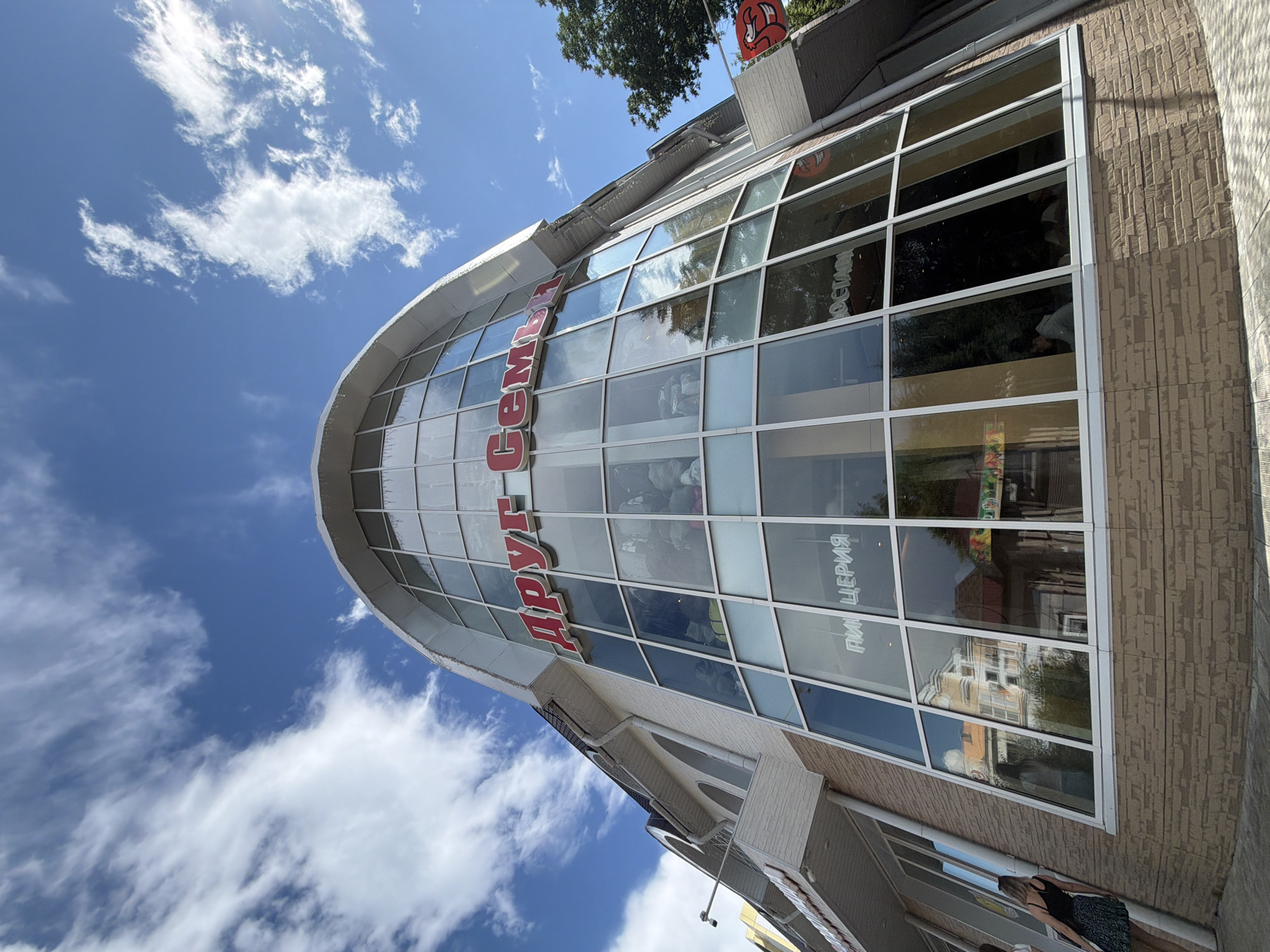 The image shows the facade of a building with a large, curved glass window. Red text is visible on the building, reading "ДРУГ СЕМЬИ" (Friend of the Family). The building appears to be a commercial establishment, possibly a store or restaurant, with products displayed behind the glass on the lower levels. Reflections of trees and the sky are visible in the windows. The sky is blue with scattered white clouds. In the foreground, a person is walking past the building. The building's facade is made of beige brick on the lower section and white siding on the upper section. The scene is set in Birobidzhan, Russia.