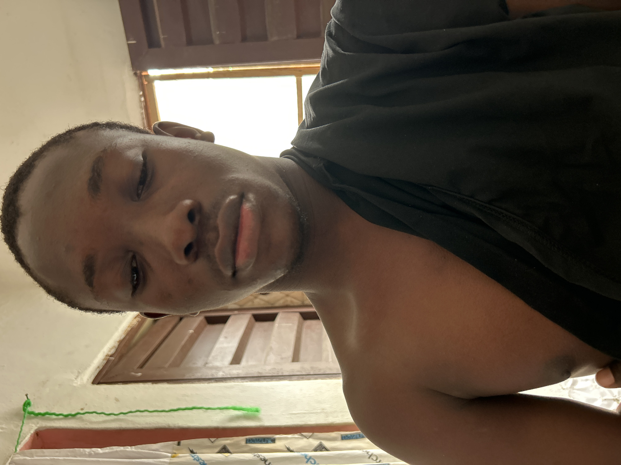The media file is a close-up, low-angle, portrait-style selfie of a young Black person. The person is wearing a black t-shirt and their face is partially visible, with their eyes closed and lips slightly parted. The background appears to be an indoor setting, possibly a room, with a window and wooden blinds. There's a hint of natural light coming from the window, suggesting it might be daytime. A strand of green yarn is visible near the bottom left of the frame, tied to what looks like a nail or hook on a light-colored wall. Below this, there are some white objects with blue and black text, possibly a mattress or bedding. The overall mood is relaxed, and the subject appears to be in a casual setting. The location context provided is Ihiagwa, Nigeria, which might suggest the type of architecture or environment visible in the background.