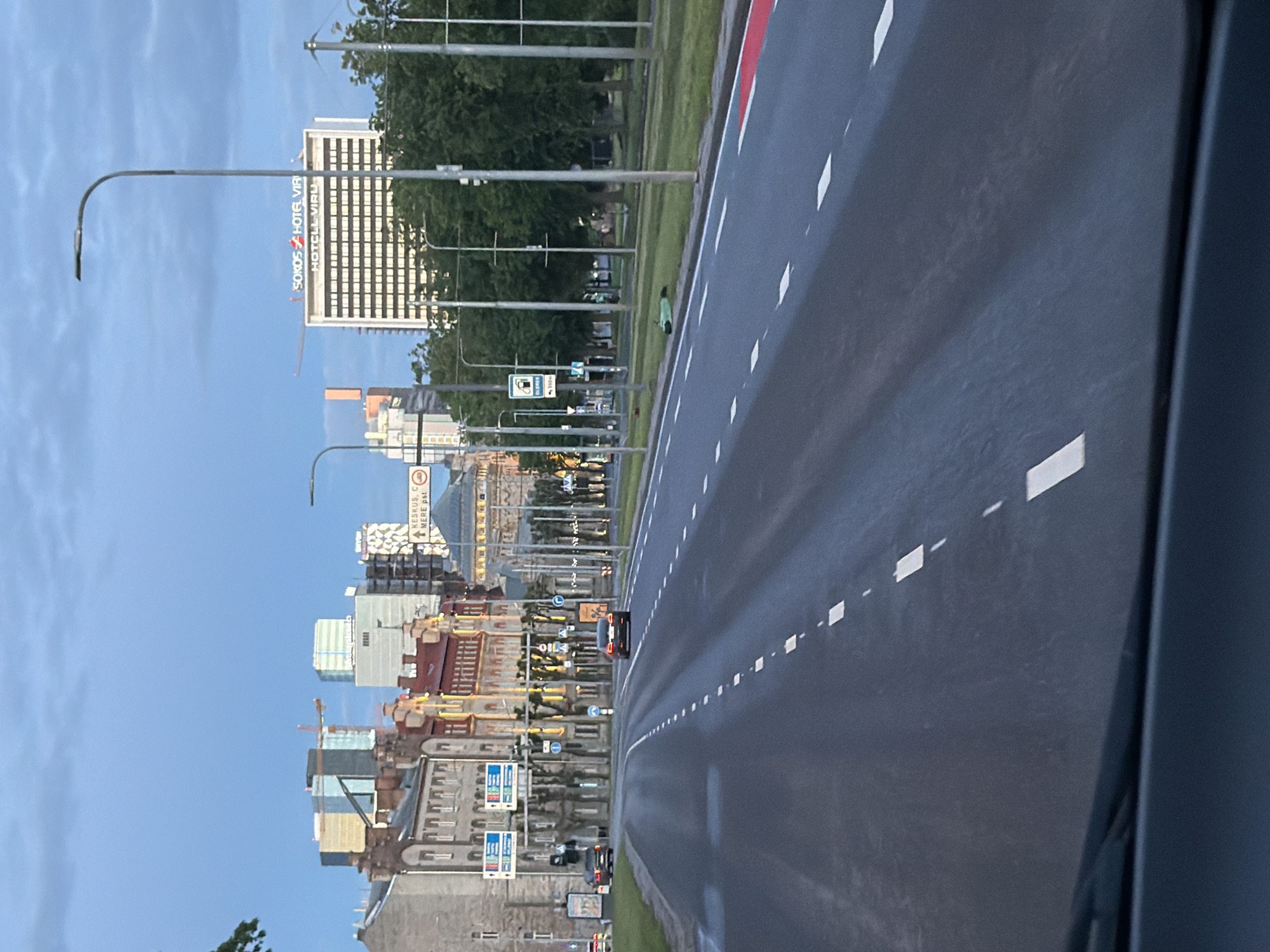 The image depicts a street scene in Tallinn, Estonia, as viewed from a vehicle. The sky is overcast with blue hues, suggesting early morning or late evening. The foreground shows the asphalt road with white dashed lane markings. In the distance, a cityscape is visible with a mix of architectural styles, including modern high-rise buildings and older, more ornate structures. One prominent building displays a sign that reads "SOKOS HOTEL VIRU". Other signs, including directional signs, are present along the street. Several cars are on the road, and trees line the sidewalks. The overall impression is that of a city thoroughfare during a period of low light.