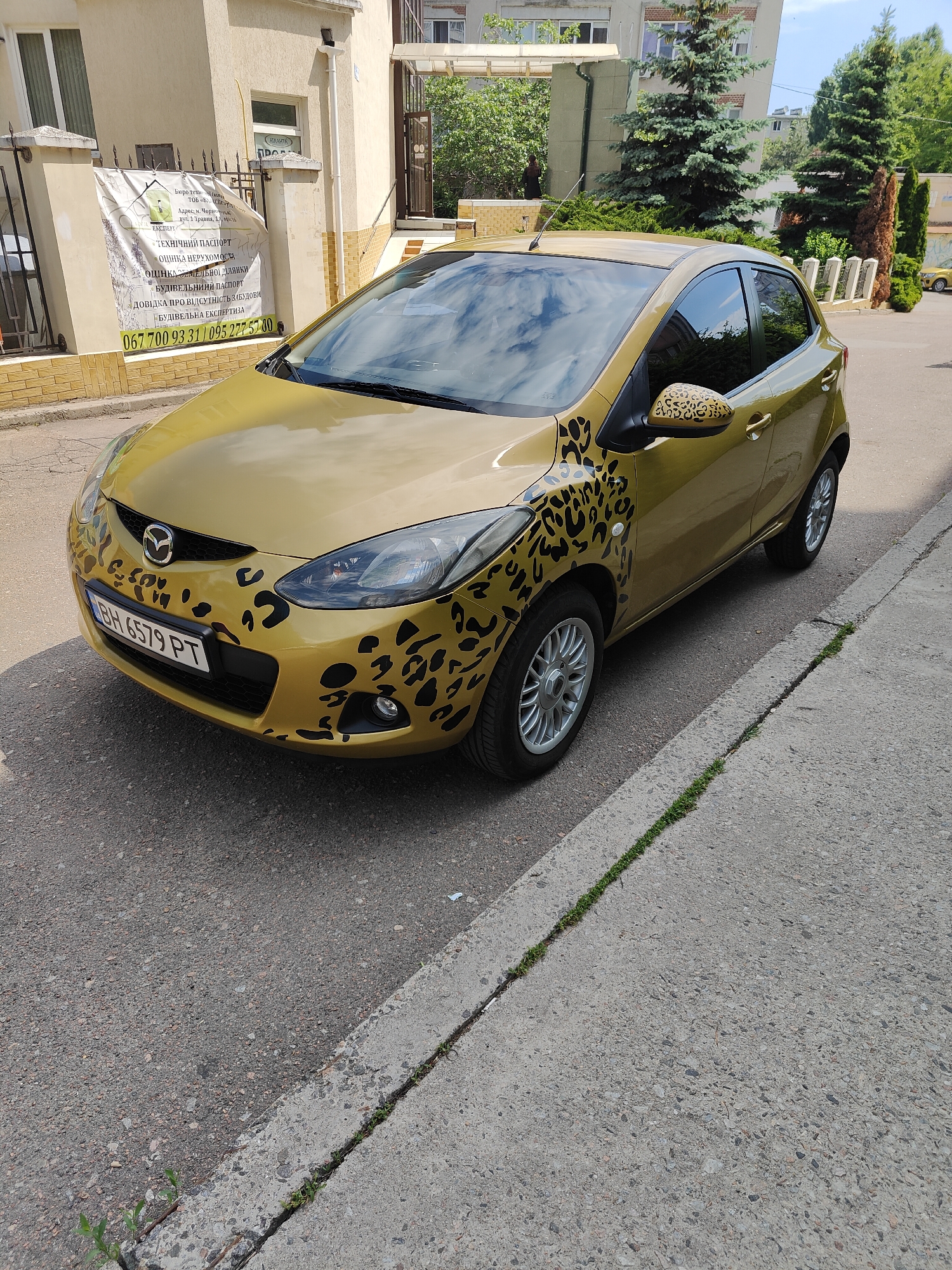 A gold-colored Mazda 2 hatchback, adorned with a leopard print decal on its front fenders and side mirror, is parked on the right side of a paved street in Chornomorsk, Ukraine. The car faces right. The street is bordered by a concrete curb and a sidewalk with sparse green grass growing in cracks. To the left of the car, a beige building with a black metal fence stands. A banner with Ukrainian text and graphics is attached to the fence. Trees and foliage are visible in the background, along with other buildings. The sky is partly cloudy. The license plate of the car reads "BH 6579 PT".
