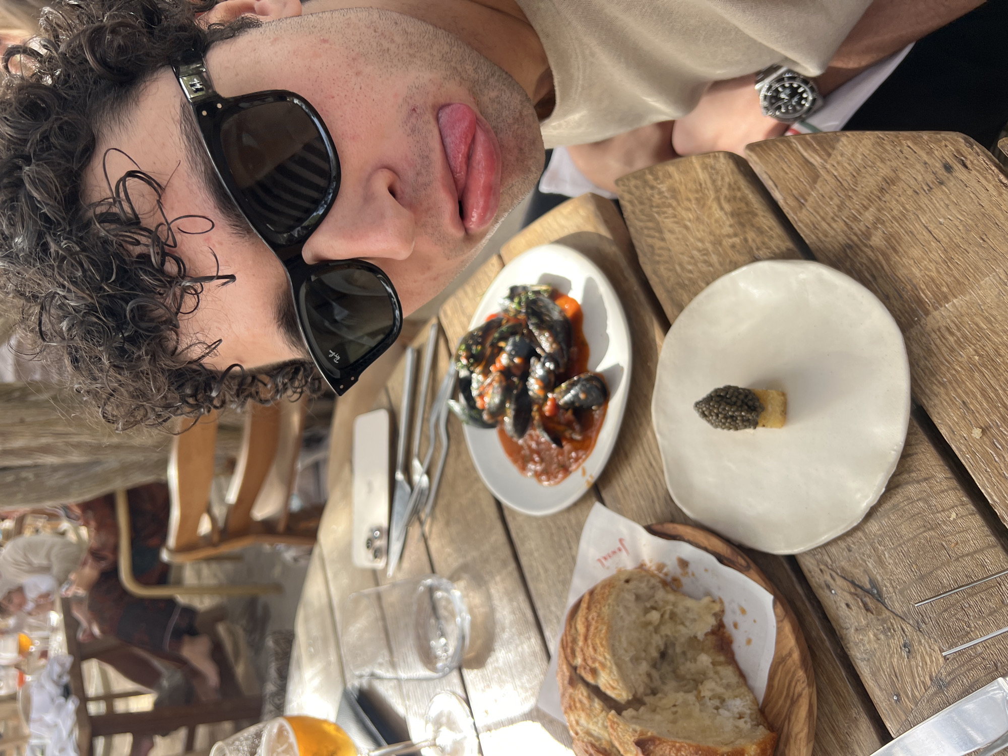 The image shows a selfie taken at a restaurant in Sant Josep de sa Talaia, Spain. A man with curly hair and sunglasses sticks his tongue out towards the camera. He wears a beige shirt and a watch is visible on his wrist. On a wooden table in front of him are several dishes, including mussels in sauce, bread, and what appears to be a small piece of food topped with caviar on a white plate. A phone, cutlery, and a glass are also on the table. In the background, other people are seated at tables, suggesting a busy restaurant atmosphere. The image is taken at an angle, giving a skewed perspective of the scene.