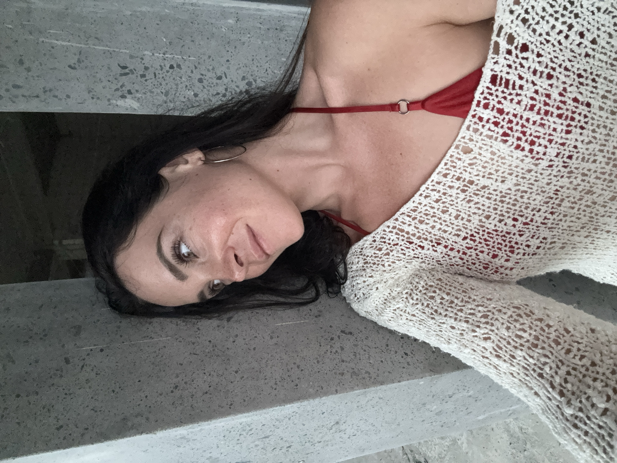 A woman with dark hair and brown eyes smiles as she looks to her left, leaning against a concrete pillar. She is wearing a red bikini top and a white, open-knit sweater draped over one shoulder. The background is dimly lit and appears to be an unfinished or industrial space with more concrete structures. The image does not provide any information about the city of Bebandem, Indonesia.