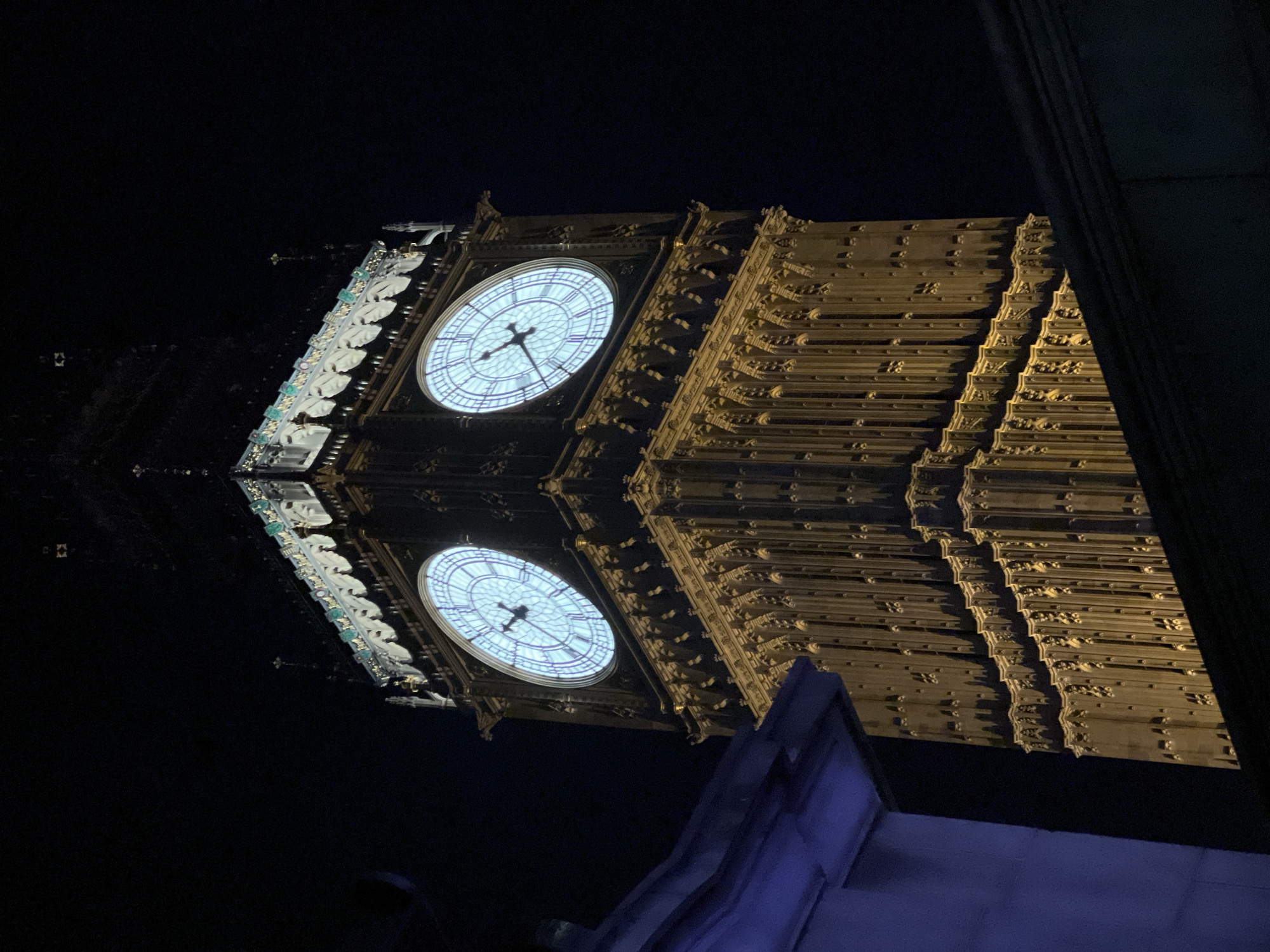 The image shows Big Ben, the clock tower at the Palace of Westminster in London, United Kingdom, at night. The clock face is illuminated. The tower appears to be reflected, creating a mirrored effect. The image includes architectural details of the tower and surrounding structures.