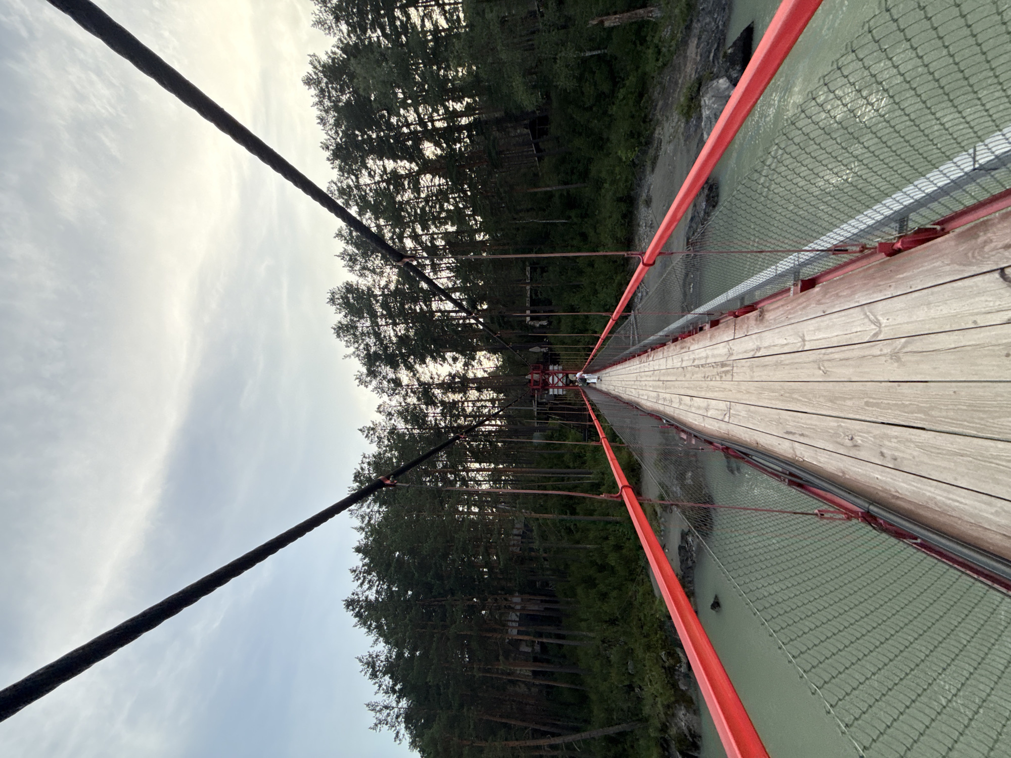 The image shows a suspension bridge spanning a river in Barangol, Russia. The bridge has a wooden walkway and is lined with red railings and a wire mesh fence. The river below is a murky green color. On the far bank of the river, a dense forest of tall pine trees rises up a hillside. The sky above is overcast with scattered clouds. In the distance, on the bridge, a person is walking towards the camera. The overall impression is one of natural beauty and a sense of remoteness.