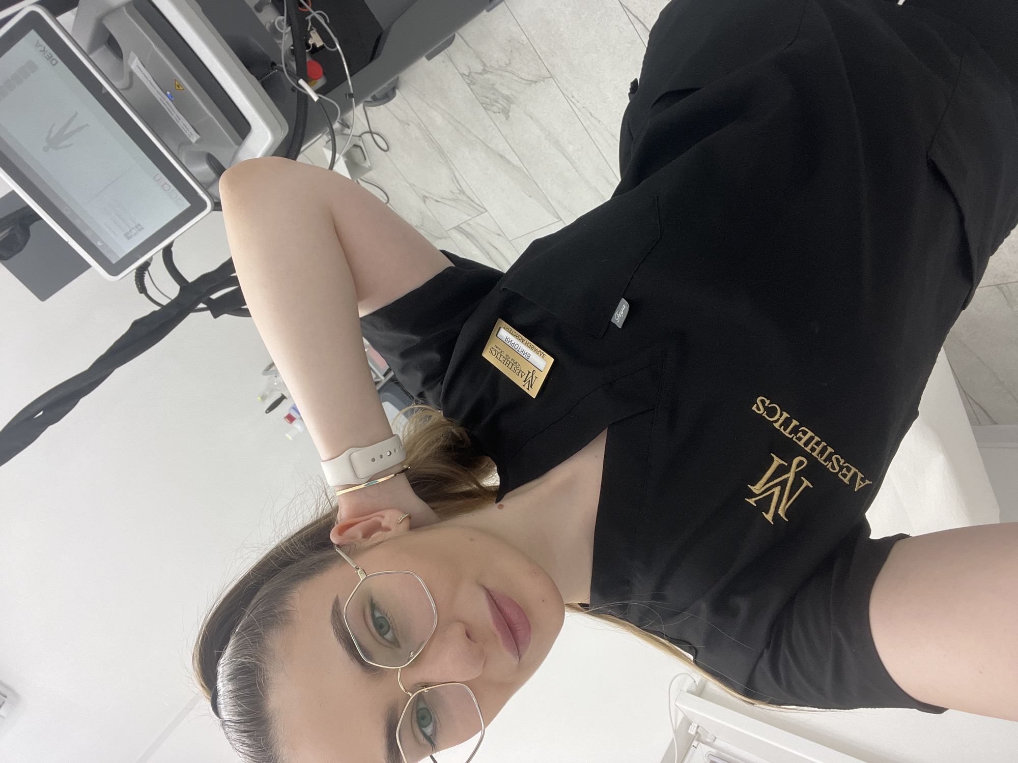 The image shows a woman with light hair, wearing glasses and a black uniform with the logo "M Aesthetics" embroidered in gold. She has a name tag that says "Виктория Заравескин", likely her name in a Cyrillic script. She appears to be in a medical or aesthetic setting, as suggested by the uniform and the medical device with a screen in the background that says "DEKA". The environment appears clean and modern with light-colored flooring, possibly tile or linoleum. No specific city is identifiable from the image.