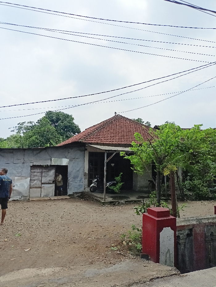 This photograph captures a rural or semi-rural scene in Ngoro, Indonesia, under an overcast sky. Numerous utility lines crisscross the sky overhead. The central focus is a house with a distinct reddish-brown tiled roof, possibly reflecting local architectural styles. To the left of the main house, an attached structure, likely an outbuilding or extension, is constructed from corrugated metal sheets.

In the foreground, the ground is unpaved dirt. On the far left, a person wearing a blue shirt and dark shorts is seen walking away from the viewer. Another individual stands in the doorway of the corrugated metal structure, looking out, wearing a light-colored shirt.

Under the porch area of the main house, a white scooter is parked. Green trees and dense foliage are visible behind and to the right of the buildings, contributing to a lush environment. In the immediate foreground on the right, there's a red and white painted concrete structure, possibly part of a low wall or a bridge railing. The scene depicts a quiet, everyday moment in a local community. No visible text is present.
