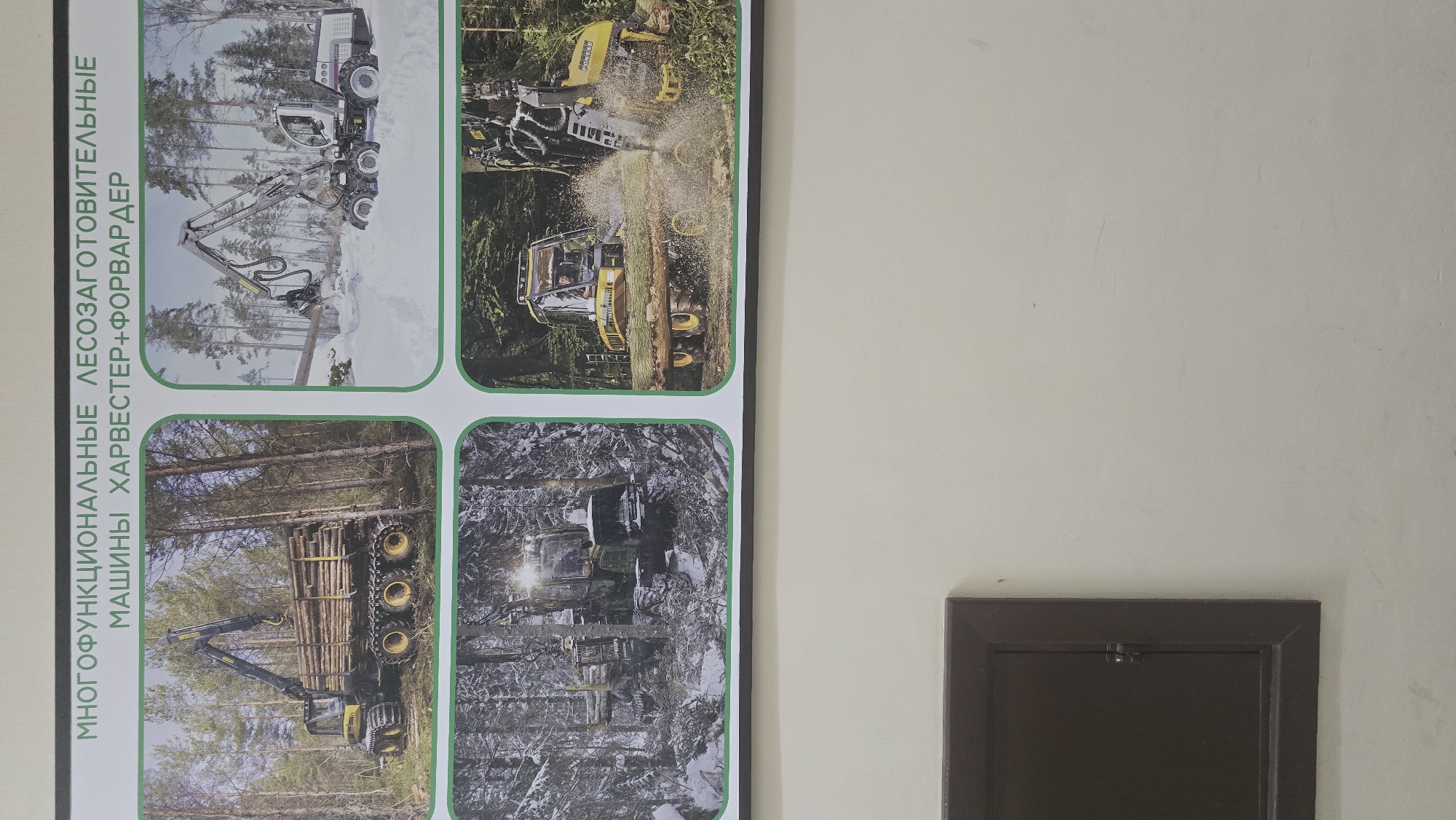 This media file is a photograph of a poster displayed on a wall, likely in a public space in Ukhta, Russia. The poster features four images of forestry equipment, specifically harvesters and forwarders. The text on the left side of the poster is in Russian and reads: "МНОГОФУНКЦИОНАЛЬНЫЕ ЛЕСОЗАГОТОВИТЕЛЬНЫЕ МАШИНЫ ХАРВЕСТЕР+ФОРВАРДЕР", which translates to "Multifunctional Logging Machines Harvester+Forwarder".
The top left image shows a white forestry machine with a large grapple arm extended, seemingly in a snowy forest. The top right image depicts a yellow forestry machine in action, cutting a tree with a chainsaw attachment, with sawdust flying. The bottom left image shows a yellow and black forwarder loaded with logs, moving through a forest. The bottom right image is a darker, more abstract view of a forestry machine in a forest, possibly covered in snow or debris, with headlights illuminated.
The overall scene is professional and informative, showcasing the capabilities of these specialized machines in a forest setting. The weather in the images appears to vary from snowy to perhaps wetter conditions. There are no people clearly visible as the main subjects, though one driver is discernible in the top right image. The poster itself is framed by a dark border. To the right of the poster, a portion of a wall and a dark, recessed panel are visible.